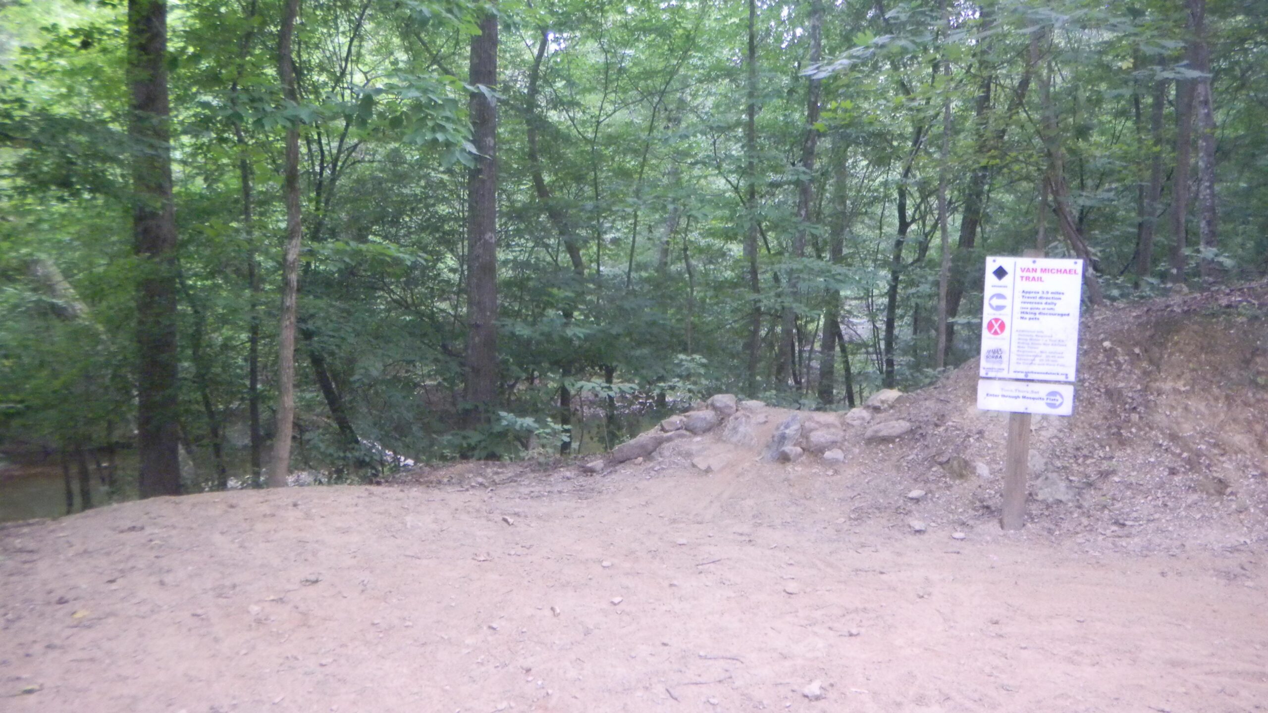 A dirt path leading into a dense forest, with a wooden sign indicating the Van Michael Trail. The sign displays trail information and guidelines, surrounded by lush green trees and a natural landscape. Blankets Creek mountain bike trail.