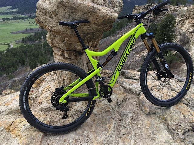 Santa Cruz Bronson: A bright green mountain bike is leaning against a large rock formation, set against a scenic landscape of rolling hills and trees in the background. The bike features thick tires, a sturdy frame, and front suspension, ready for off-road adventures.