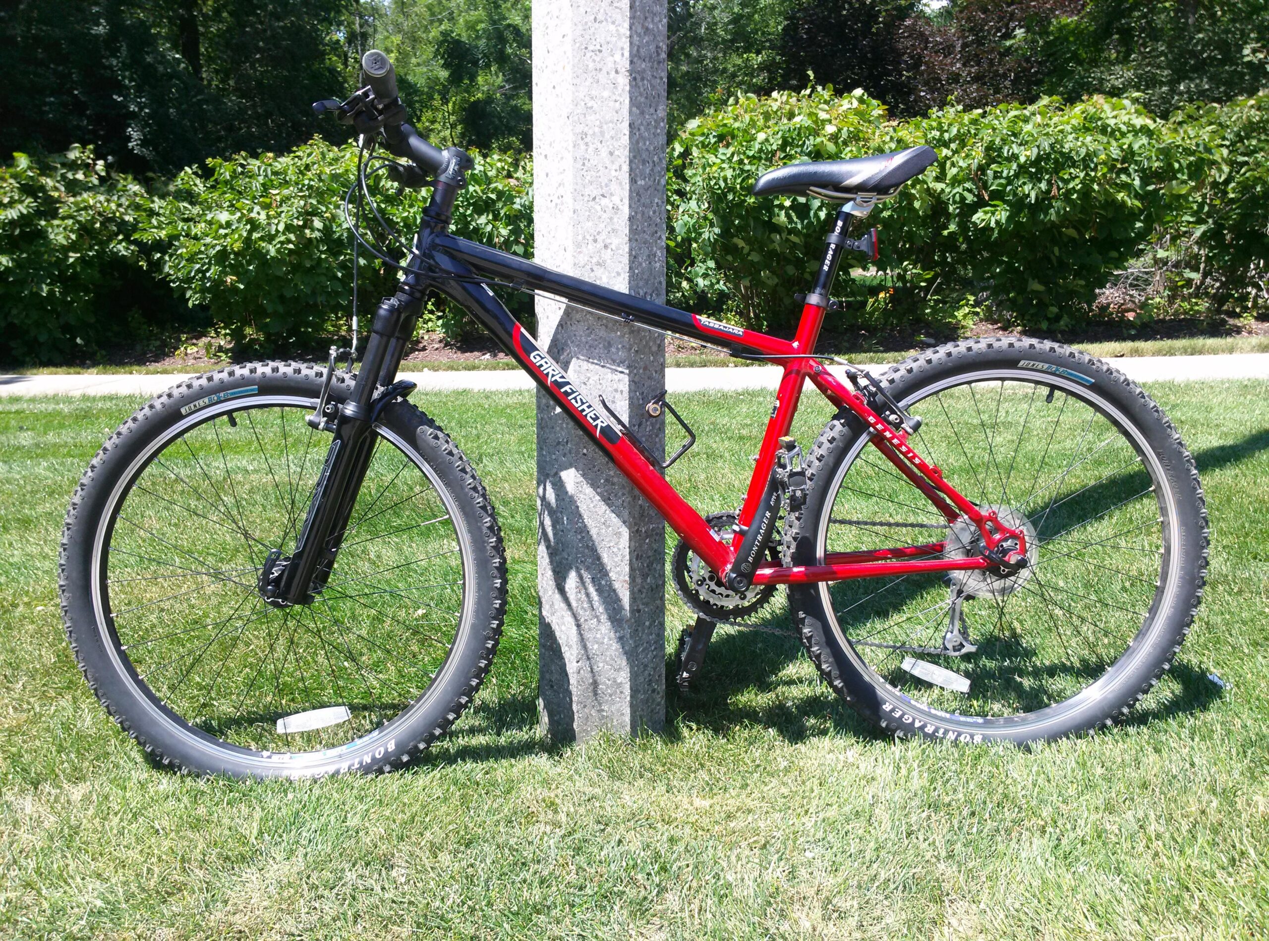 Gary Fisher Tassajara: A red and black mountain bike leaning against a stone pole, set against a grassy background with bushes. The bike features thick tires designed for off-road use and a textured handlebar grip.