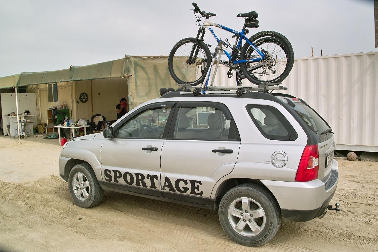 Giant Yukon: A silver SUV with the word "SPORTAGE" printed on its side, parked on sandy ground. A blue mountain bike is secured on the roof rack. In the background, a makeshift shelter with a few people and various items is visible.