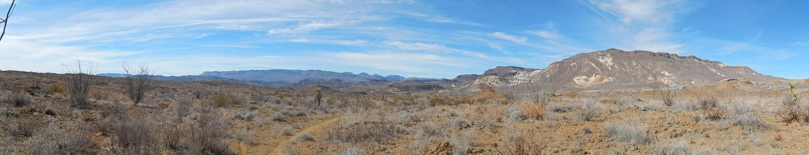 A panoramic view of a vast desert landscape under a clear blue sky. The scene features gently rolling hills and distant mountains, with sparse vegetation and dry, sandy ground. The environment conveys a sense of arid beauty, typical of a desert region. Big Bend State Park mountain bike trail.