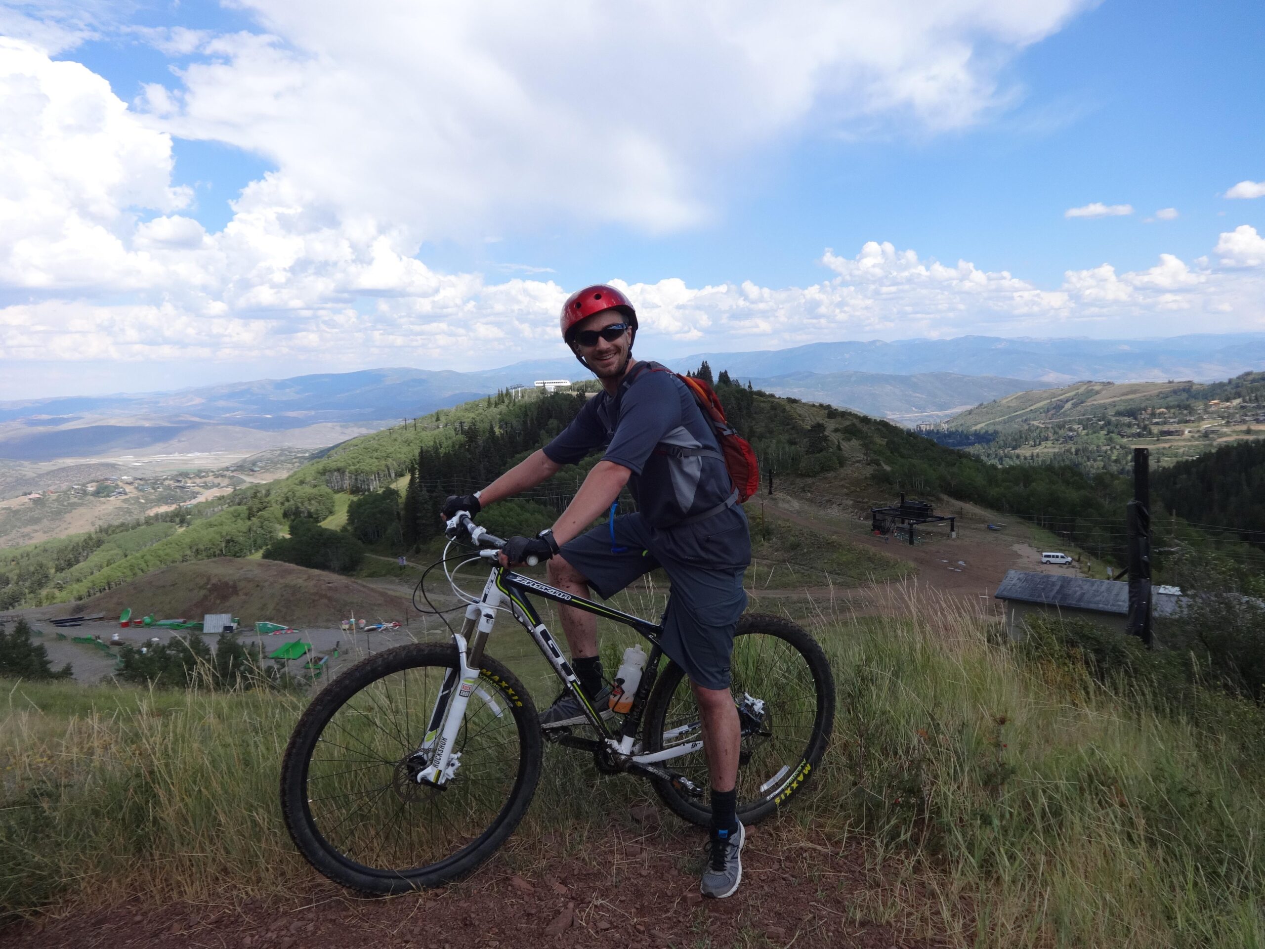A person wearing a helmet and sunglasses is posing next to a mountain bike on a grassy hillside, with a scenic view of mountains and a valley in the background under a partly cloudy sky. Mid Mountain mountain bike trail.