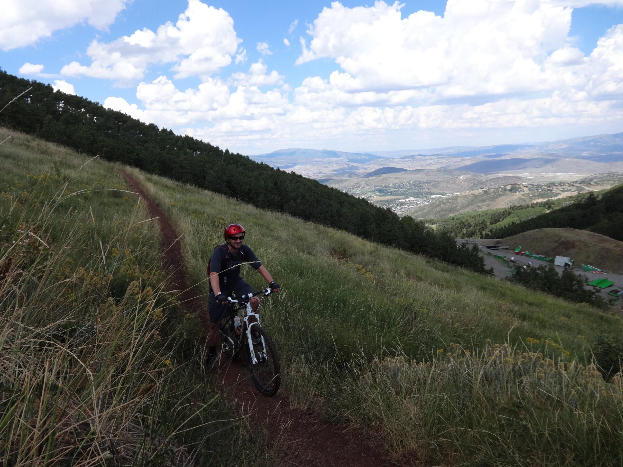 A mountain biker rides along a narrow dirt trail, surrounded by tall grass and trees on a hillside. The backdrop features a panoramic view of a valley and distant mountains under a partly cloudy sky. Mid Mountain mountain bike trail.