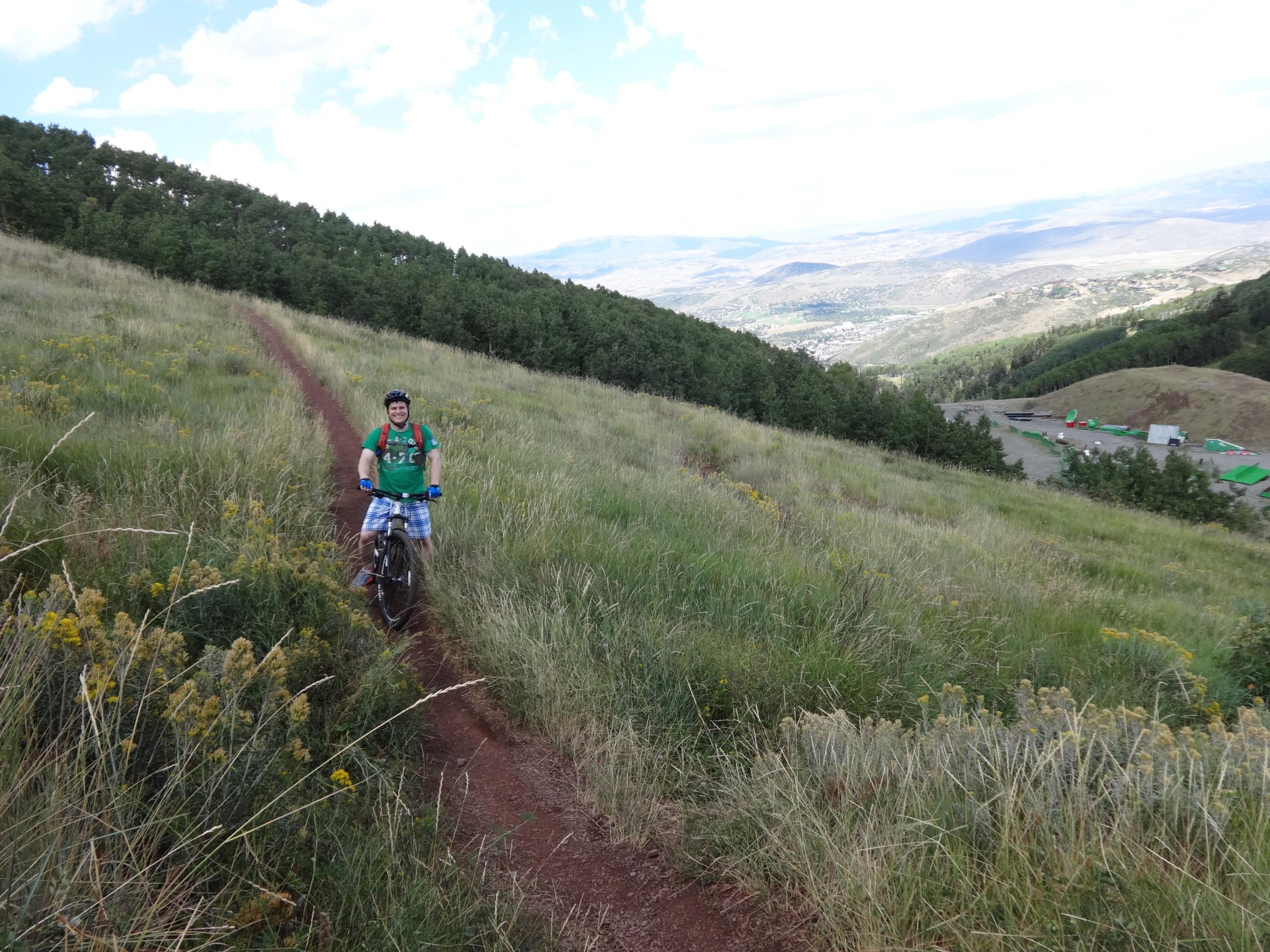 A person riding a mountain bike on a dirt trail surrounded by tall grass and wildflowers, with a backdrop of green trees and a distant view of hills and valleys under a partly cloudy sky. Mid Mountain mountain bike trail.
