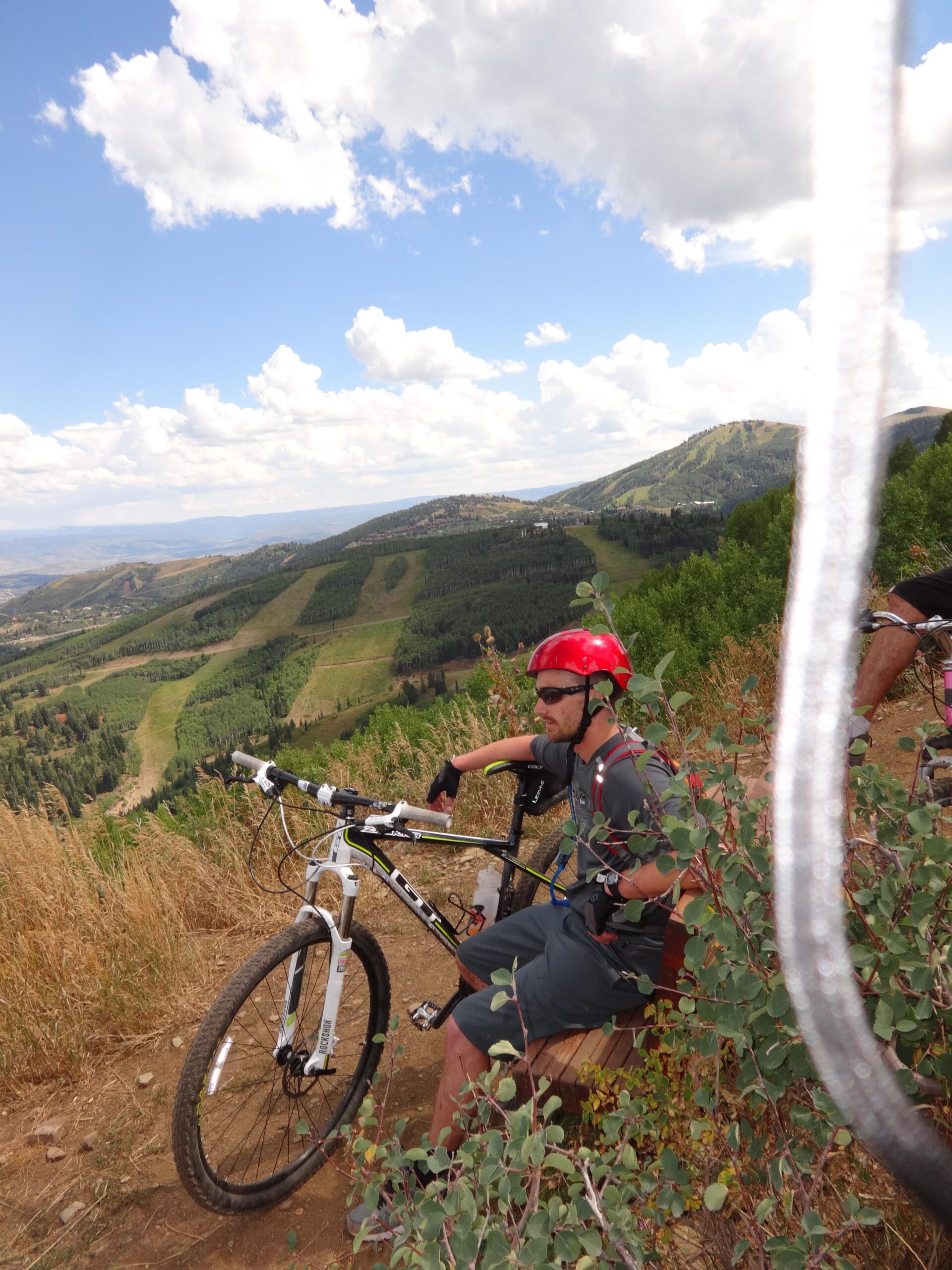 A mountain biker in a red helmet and sunglasses sits on a wooden bench, resting while leaning against his bike. He is surrounded by tall grass and trees, with a scenic view of rolling hills and a cloudy blue sky in the background. Mid Mountain mountain bike trail.