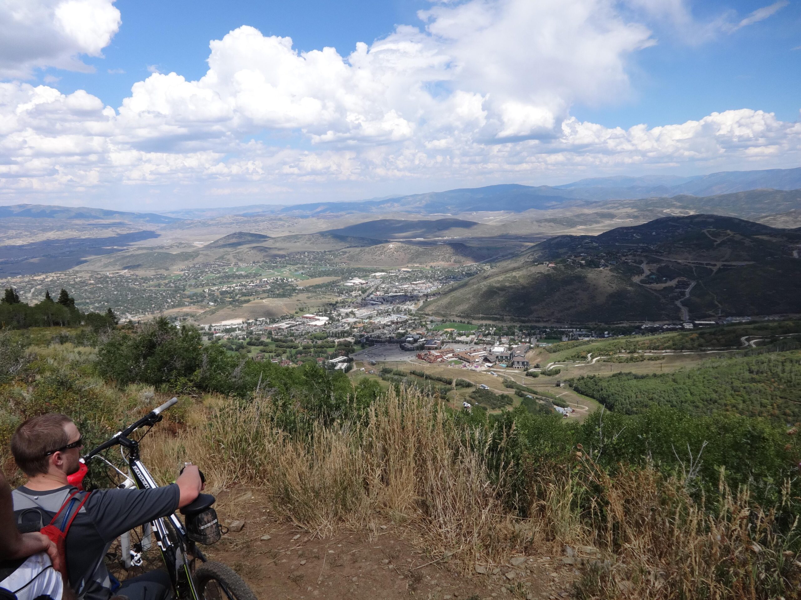 A mountain biker sitting on a hillside, overlooking a valley and a small town surrounded by green landscaping and rolling hills. The sky is partly cloudy, with blue and white hues creating a scenic backdrop. The biker's bicycle is resting beside him as he takes in the view. Mid Mountain mountain bike trail.