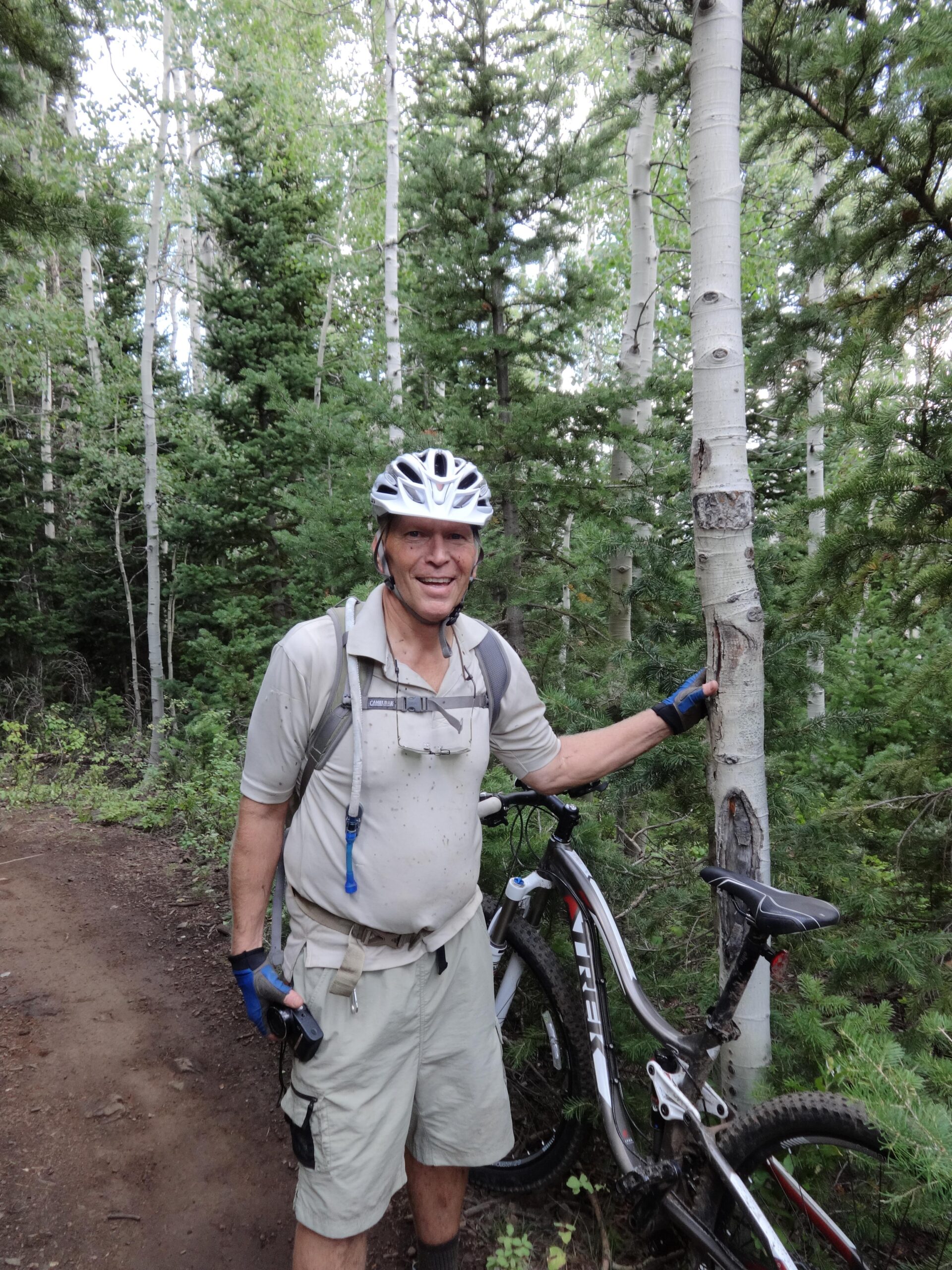 A person wearing a bicycle helmet and gloves stands next to a mountain bike on a dirt trail surrounded by tall trees. The individual is smiling and appears to be taking a break in a forested area. Mid Mountain mountain bike trail.