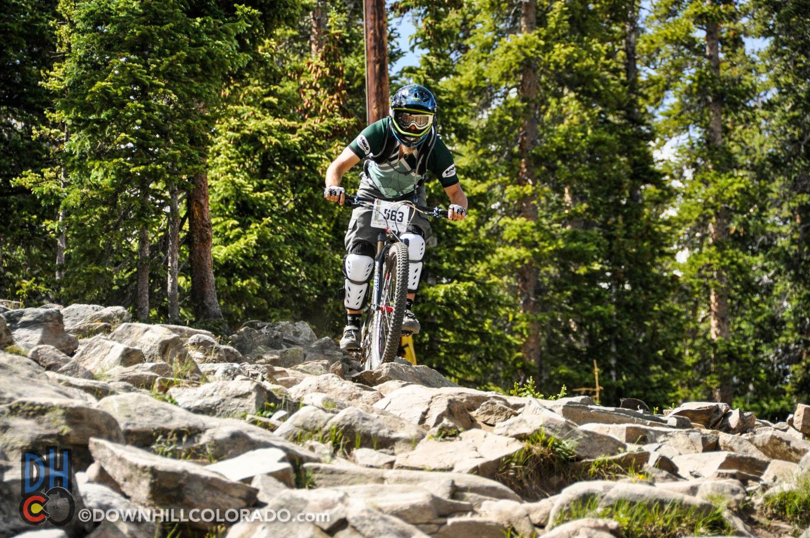 A mountain biker navigating rocky terrain in a forested area, wearing protective gear and a helmet. The biker is in motion, showcasing an action shot of the descent on a challenging trail. Keystone Resort Bike Park mountain bike trail.