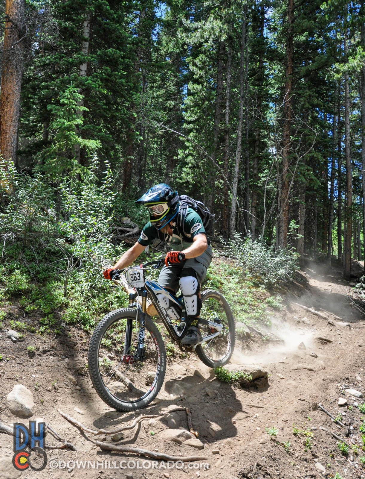 A mountain biker navigating a dirt trail in a pine forest, kicking up dust, wearing a helmet and protective gear. The sunlight filters through the trees, highlighting the rider's focused expression and dynamic stance as they maneuver around a curve. Keystone Resort Bike Park mountain bike trail.