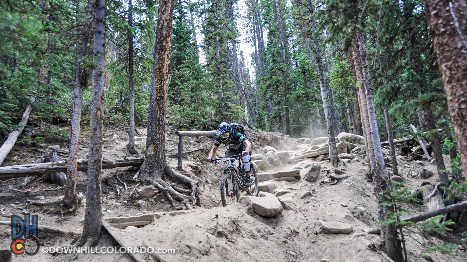 A mountain biker navigates a rocky trail through a dense forest, with dust kicked up from the terrain. Tall trees surround the path, creating a natural setting for off-road cycling. The biker is equipped with safety gear, including a helmet and knee pads. Keystone Resort Bike Park mountain bike trail.