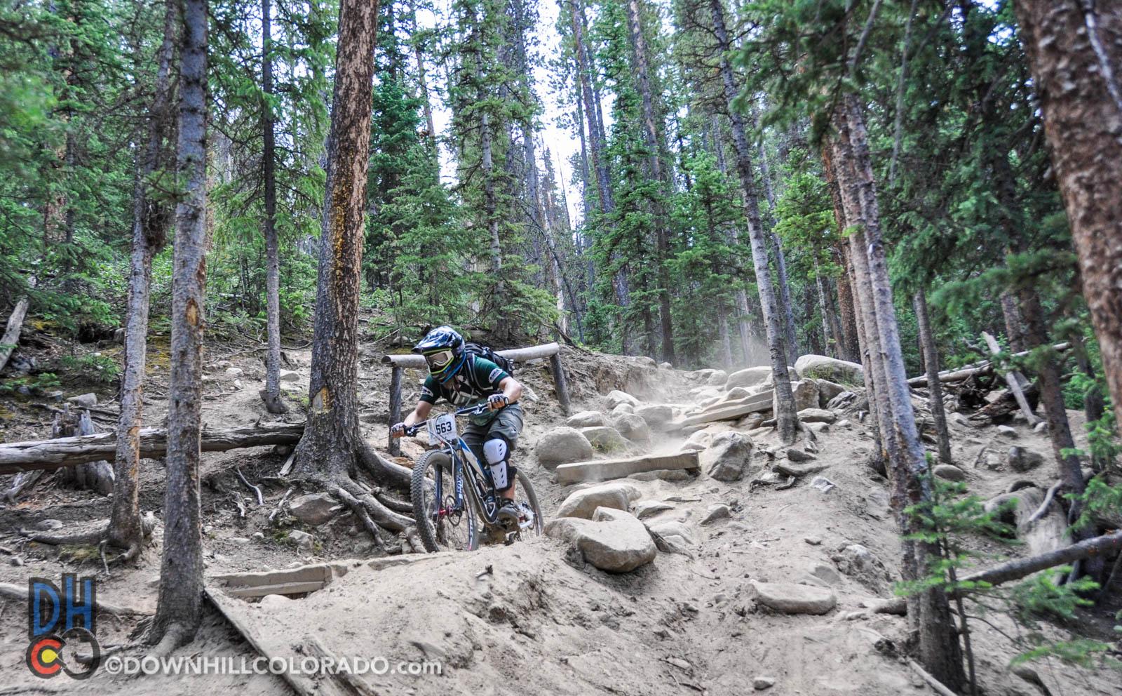 A mountain biker navigating a rocky, dusty trail through a dense forest of tall trees. The biker, wearing protective gear and colored clothing, is focused on maneuvering over the uneven terrain, with dust swirling around from the bike's movement. Keystone Resort Bike Park mountain bike trail.