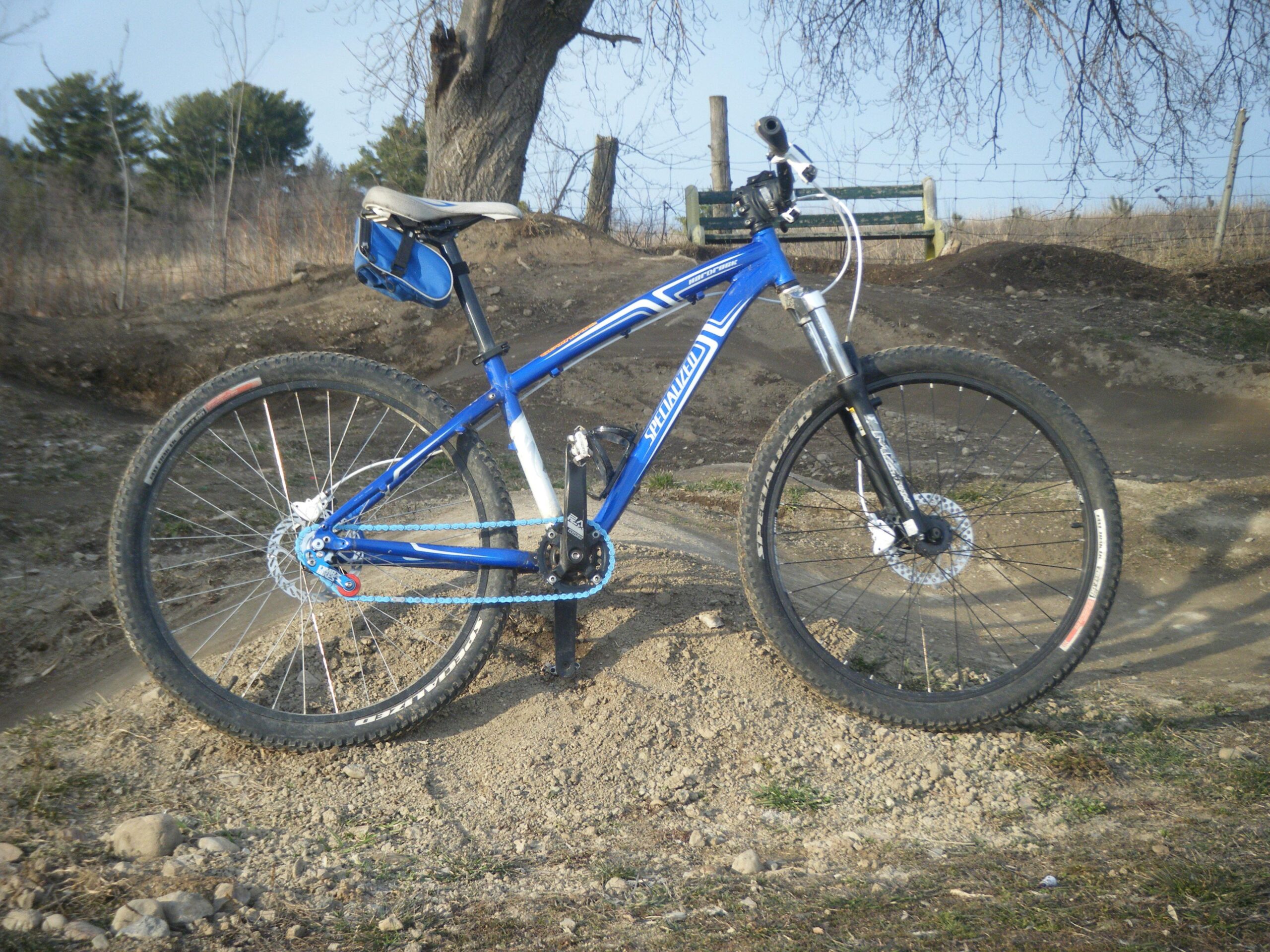 Specialized Hardrock Disc: A blue mountain bike parked on a dirt trail, with a gravel and grass surface in the background. There is a tree and a wooden bench in the distance, along with a fence line. The bike features thick tires designed for off-road riding and has a small blue bag attached to the seat.