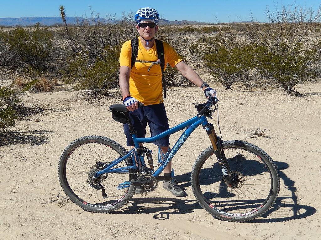A person wearing a yellow shirt, blue shorts, and a helmet stands next to a blue mountain bike on a sandy trail, surrounded by sparse desert vegetation under a clear blue sky. Big Bend Ranch State Park mountain bike trail.