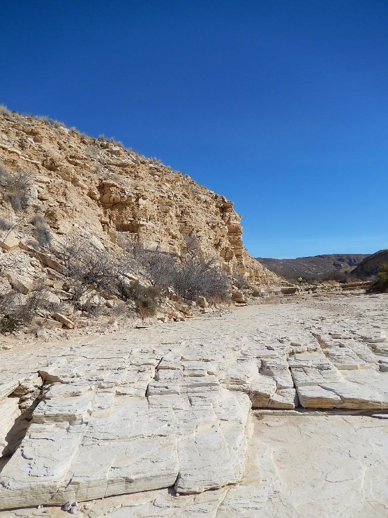 A dry, rocky landscape featuring steep, light-colored cliffs on one side, with a flat, cracked surface and sparse vegetation. The sky is clear and bright blue, suggesting a sunny day in a desert or arid region. Big Bend Ranch State Park mountain bike trail.