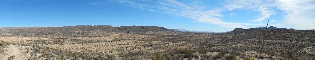 A panoramic view of a desert landscape featuring rugged mountains under a clear blue sky. The foreground consists of arid terrain with sparse vegetation and rocky formations, extending into the distance where bolder mountain ranges can be seen on the horizon. Big Bend State Park mountain bike trail.