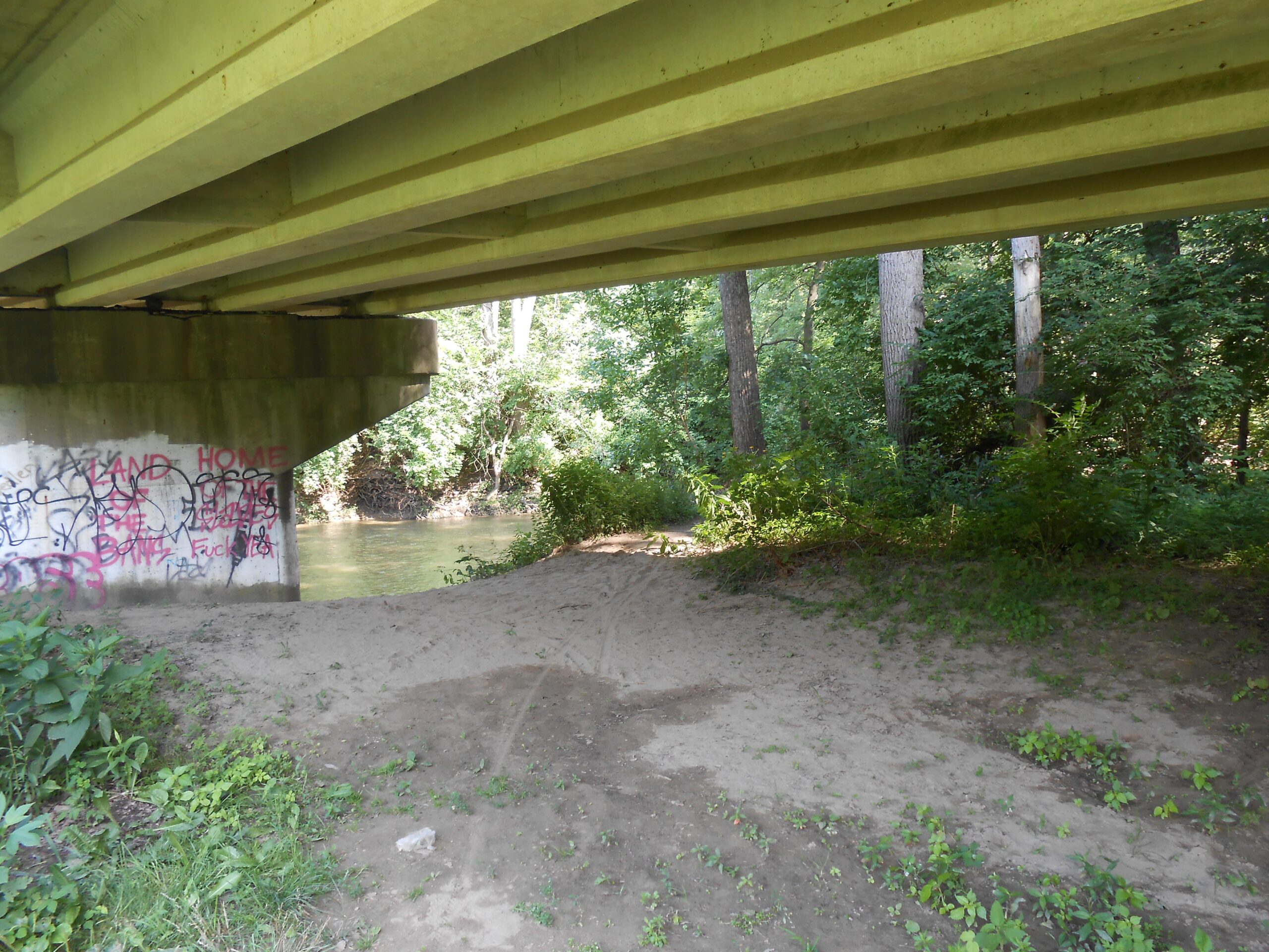 Under a bridge, with a concrete support partially covered in graffiti, a sandy area leads to a river surrounded by lush green trees. The scene captures the tranquility of nature juxtaposed with urban art. Water George mountain bike trail.
