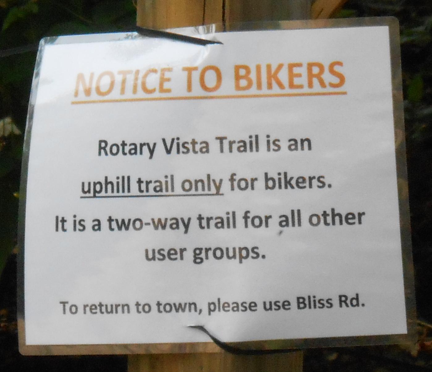 A close-up of a sign attached to a wooden post, providing important information for trail users. The sign states "NOTICE TO BIKERS" and informs that the Rotary Vista Trail is an uphill trail designated only for bikers, while it is a two-way trail for all other user groups. It also gives directions to return to town via Bliss Rd.  Rotary Vista mountain bike trail.