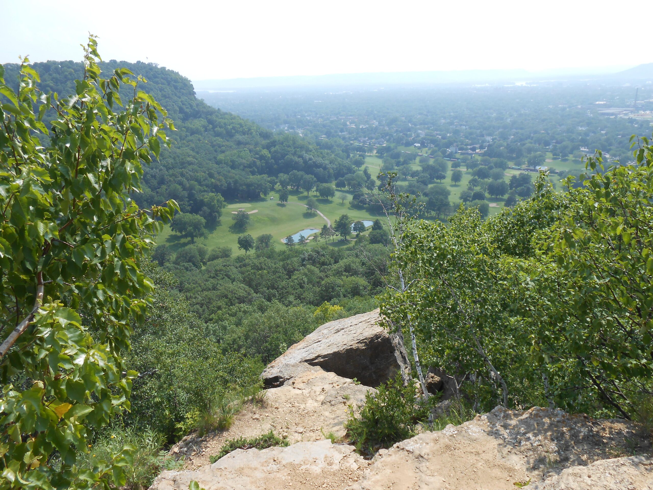 A scenic view from a rocky overlook, showing lush green hills and a valley below with a golf course and trees. The sky is hazy, suggesting a warm, sunny day. Rotary Vista mountain bike trail.
