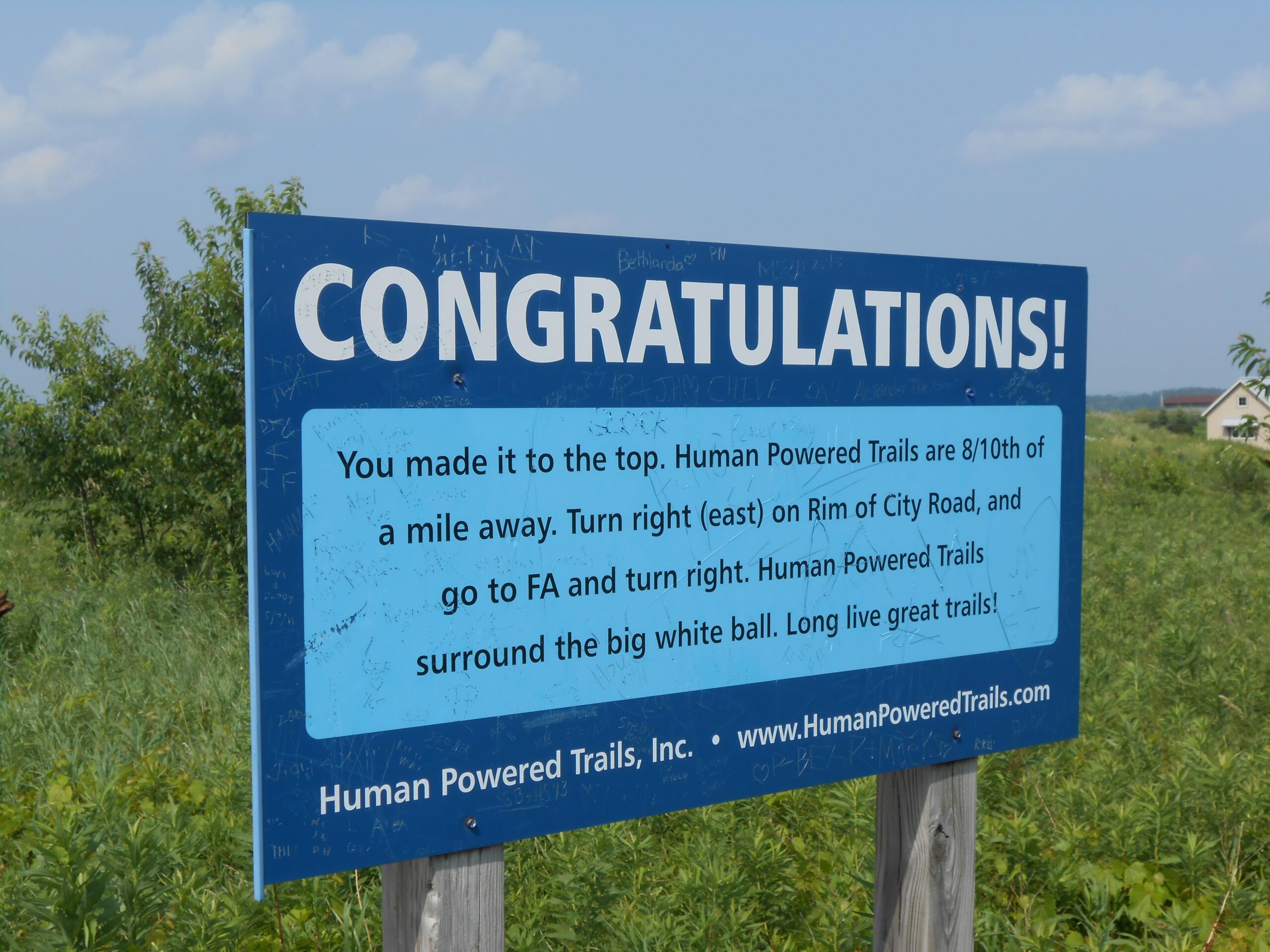 A blue sign with white text that reads "CONGRATULATIONS!" at the top, followed by instructions indicating that visitors have reached the top and detailing directions to the Human Powered Trails. The sign indicates that the trails are 8/10 of a mile away and provides guidance on turning right at Rim of City Road. The background features green vegetation and a blue sky. Rotary Vista mountain bike trail.