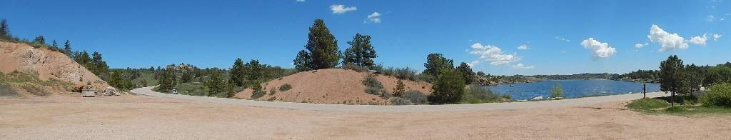 Panoramic view of a serene outdoor landscape featuring a dirt road next to a calm lake, surrounded by green hills and trees under a clear blue sky with some white clouds. Curt Gowdy State Park mountain bike trail.