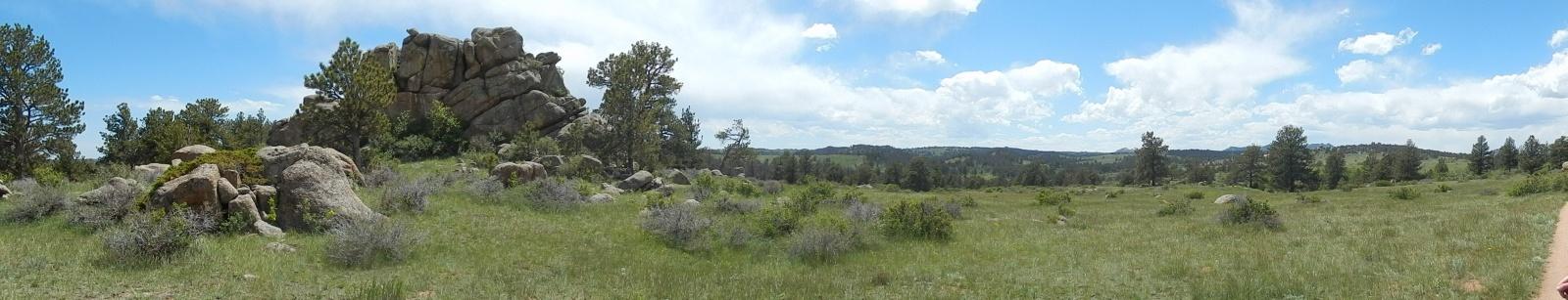 A panoramic view of a natural landscape featuring large rock formations and a grassy meadow, surrounded by trees under a partly cloudy sky. The scene showcases the beauty of the outdoors with rolling hills in the background. Curt Gowdy State Park mountain bike trail.