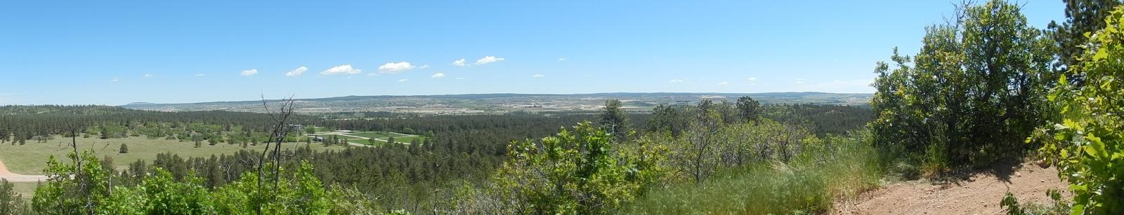 A panoramic view of a lush, green landscape featuring rolling hills and sparse trees under a clear blue sky with a few fluffy clouds. The foreground includes vibrant greenery with shrubs and trees, while the background reveals a vast expanse of more forested area and distant hills. Falcon Trail mountain bike trail.