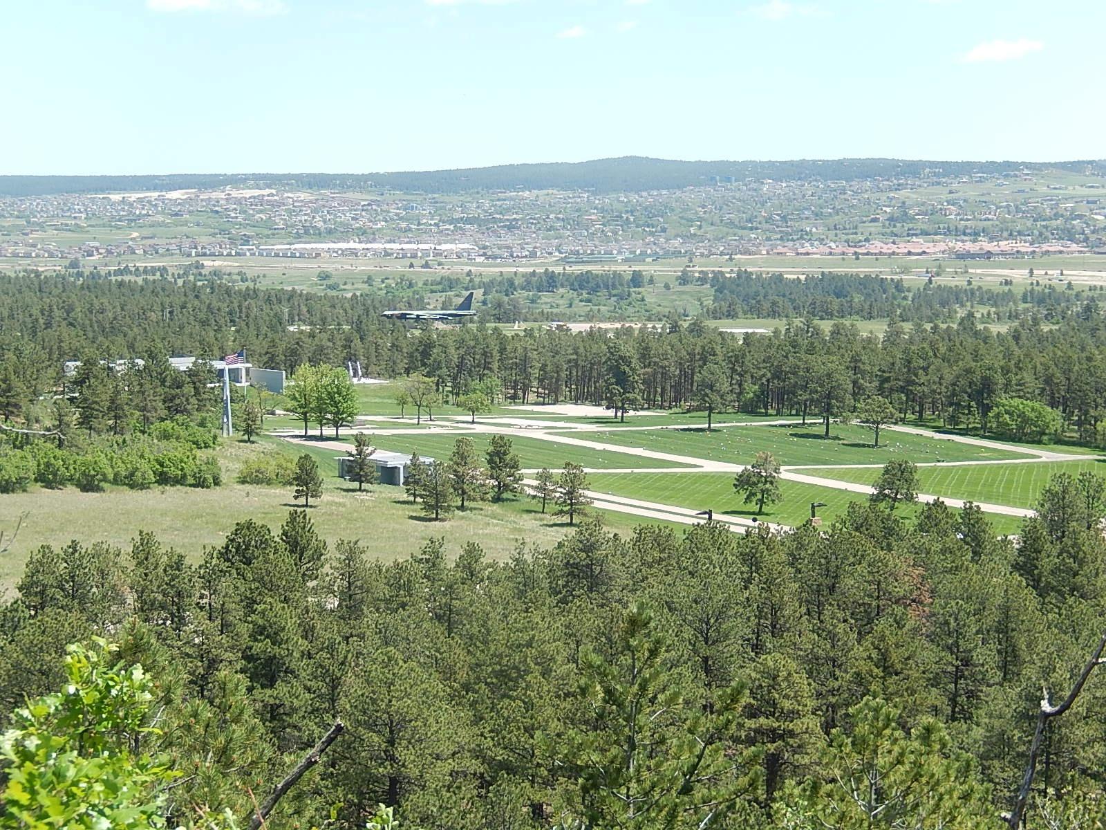 A scenic view of a landscaped area with neatly arranged greenery and roads, framed by dense trees. In the background, a small aircraft is flying overhead, while a town and rolling hills are visible in the distance under a clear blue sky. Falcon Trail mountain bike trail.