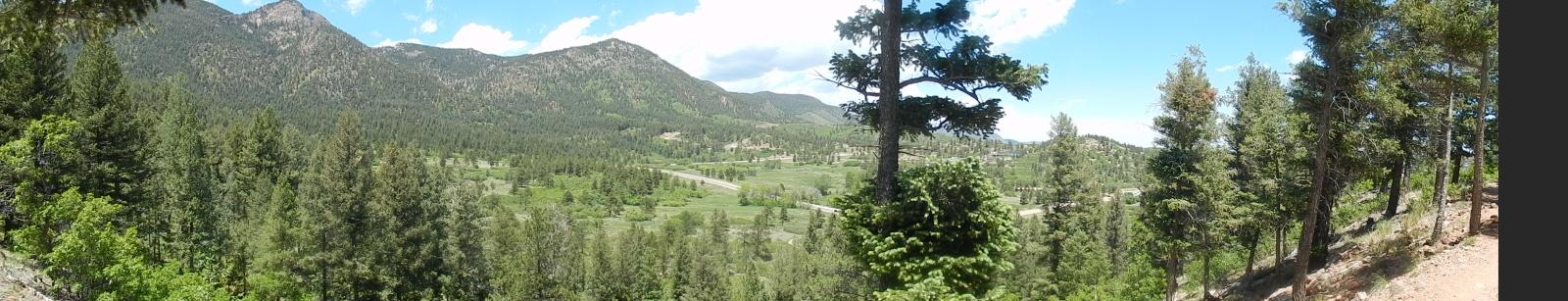A panoramic view of a lush green valley surrounded by mountains under a partly cloudy sky. The foreground features tall coniferous trees, while the background displays rolling hills and distant peaks, creating a serene natural landscape. Falcon Trail mountain bike trail.