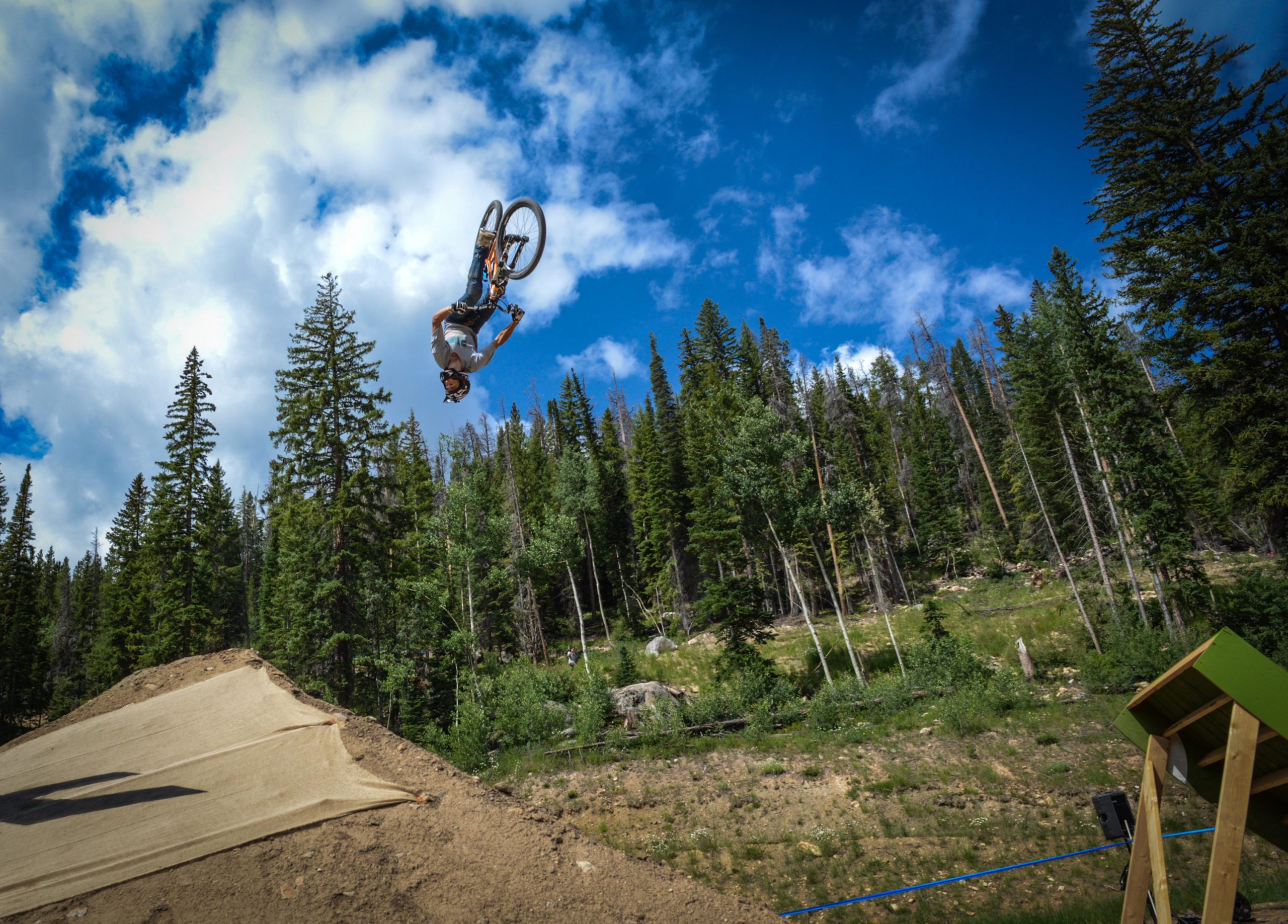 A mountain biker performs a backflip in mid-air above a dirt jump, surrounded by lush green trees and a clear blue sky with scattered clouds. Trestle Bike Park mountain bike trail.