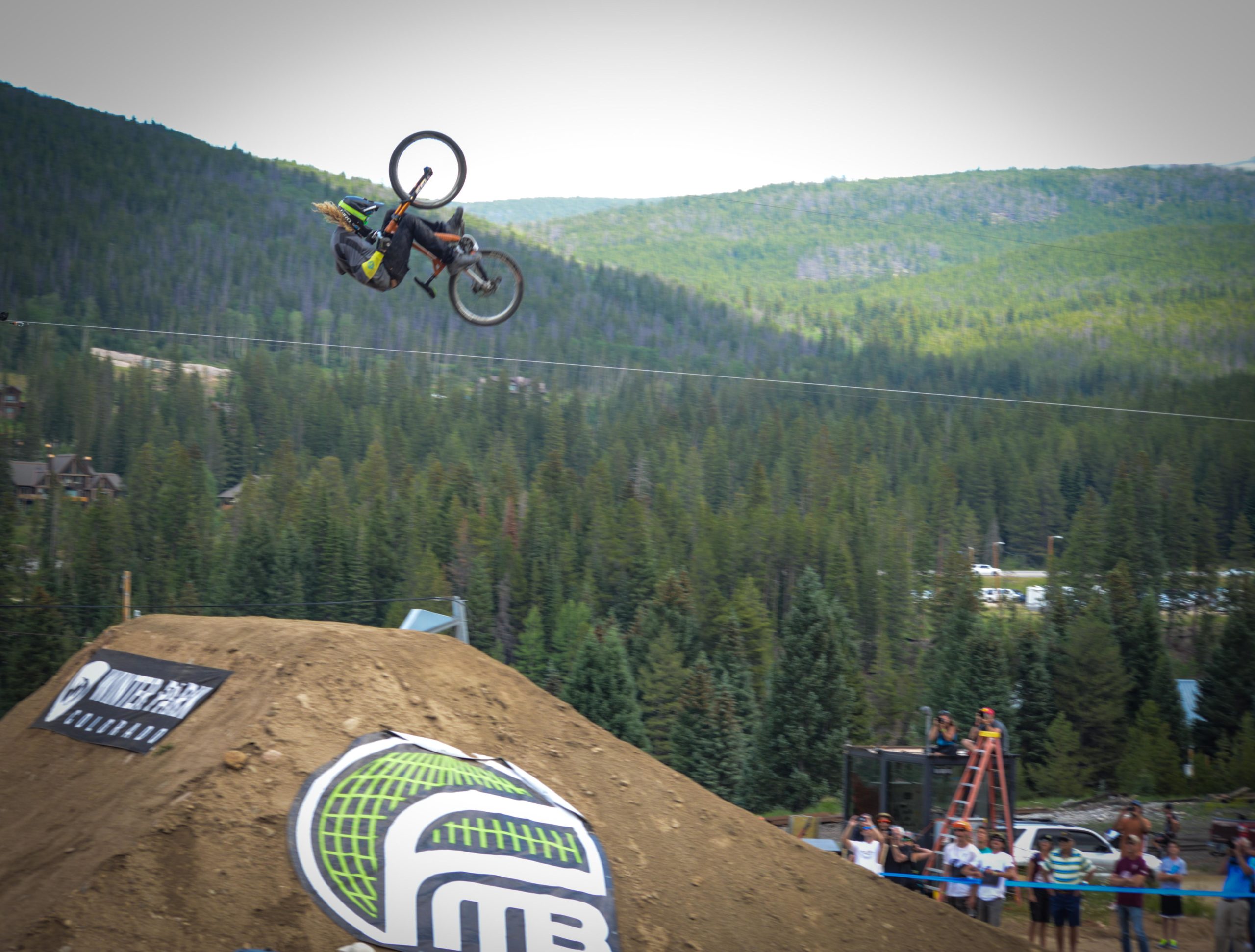 A mountain biker performing a backflip over a large dirt jump at a bike competition, with a backdrop of lush green trees and a crowd watching in the foreground. Trestle Bike Park mountain bike trail.