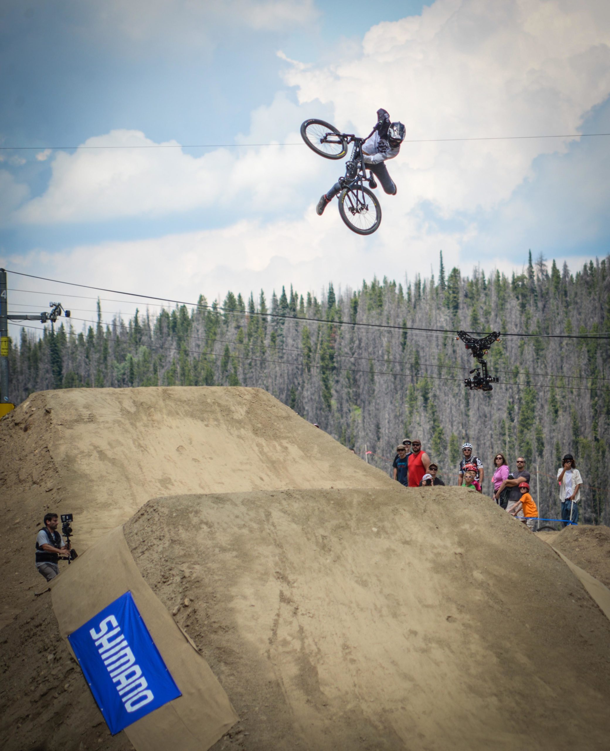 A mountain biker performs a jump over a dirt ramp while upside down, showcasing impressive aerial skills. In the background, spectators watch the event, and a camera drone hovers nearby. A blue Shimano banner is visible on the ground, adding context to the scene. The setting features trees and a blue sky with clouds. Trestle Bike Park mountain bike trail.
