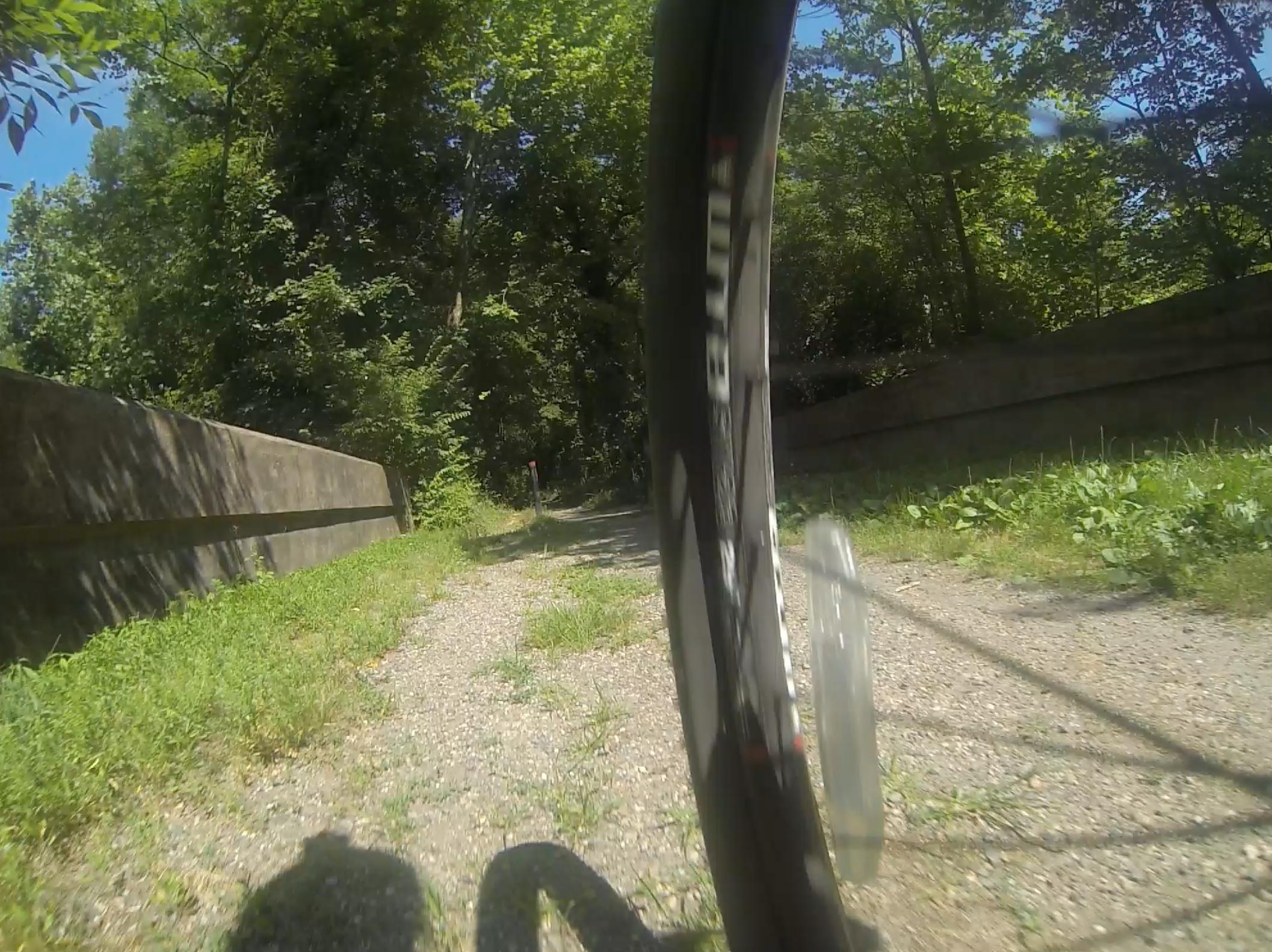 A close-up view of a bicycle wheel on a gravel path surrounded by lush greenery, with a concrete barrier on the left side. Sunlight filters through the trees, creating a bright and inviting atmosphere for outdoor biking. Difficult Run mountain bike trail.