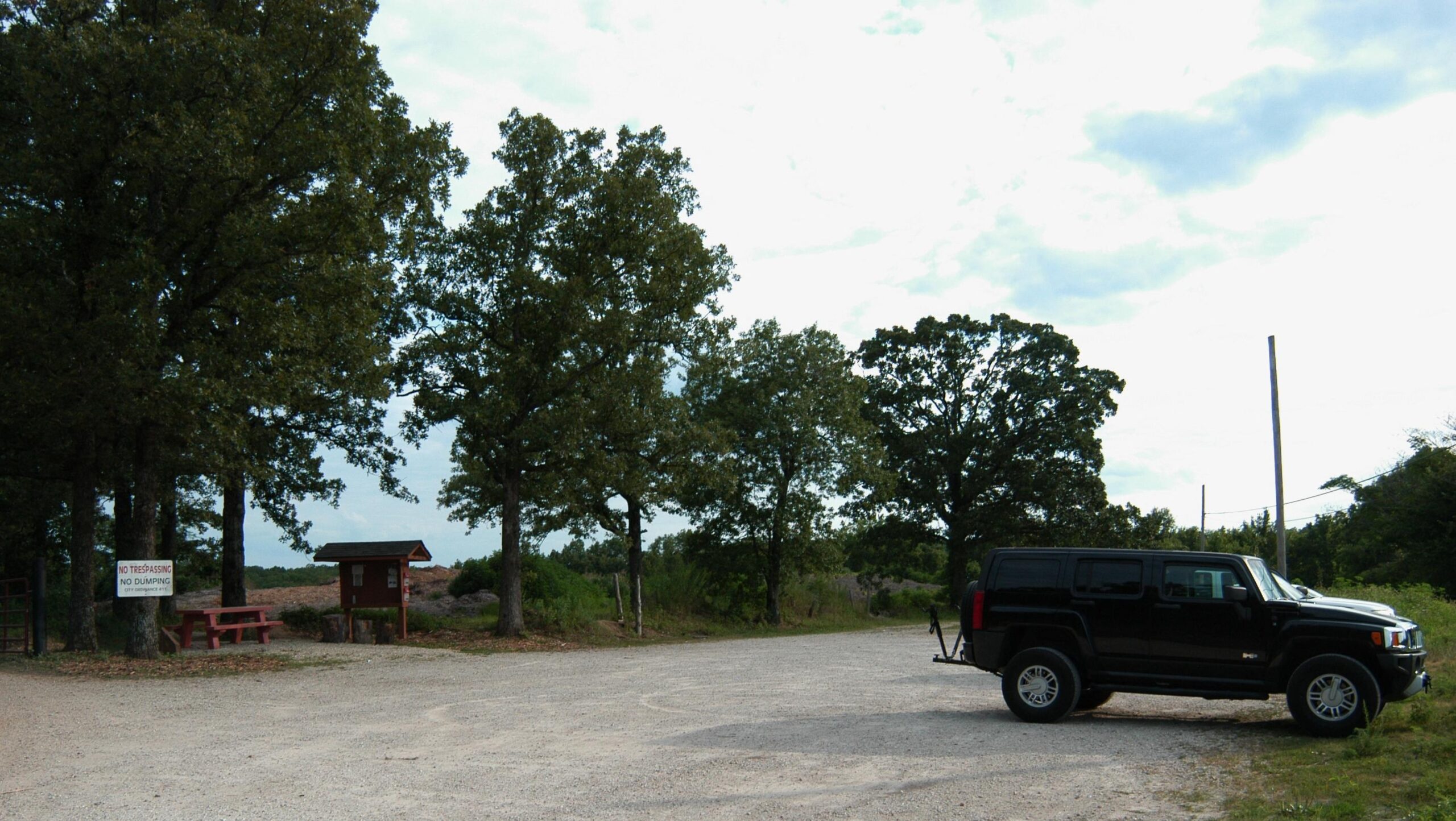 A gravel parking area surrounded by trees, featuring a black SUV parked to the right. In the background, a small wooden information kiosk is visible next to a picnic table. A sign is posted nearby, indicating "No Trespassing" and "No Dumping." The sky is partly cloudy, suggesting an overcast day. Forest City Trail mountain bike trail.