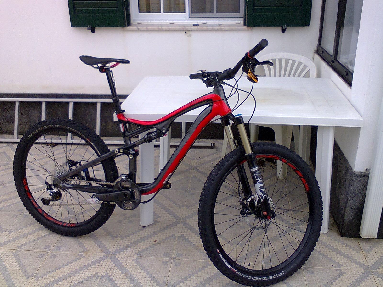 Specialized Stumpjumper FSR Elite: A red and black mountain bike parked next to a white table and chair on a patterned tiled surface, with a window and green shutters visible in the background.