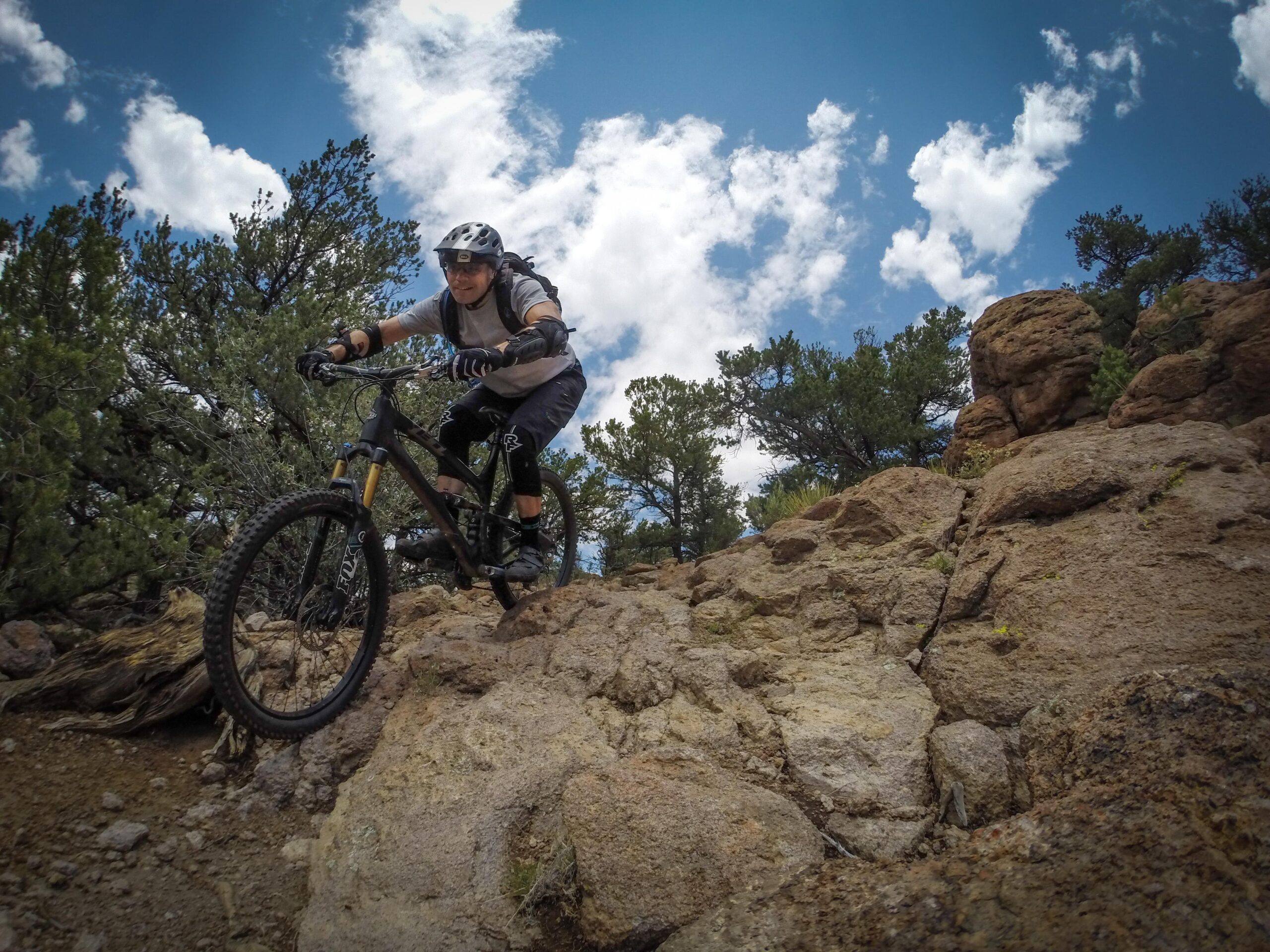 A mountain biker navigating rocky terrain with trees and a blue sky in the background. The biker is wearing a helmet and protective gear, showing focus as they descend a rocky path. Unkle Nazty mountain bike trail.