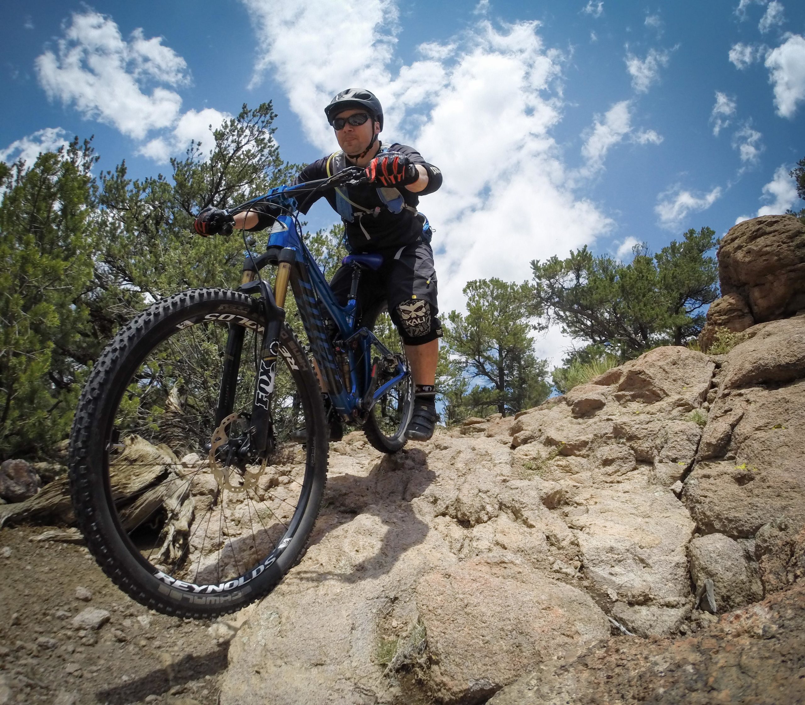A mountain biker riding on rocky terrain under a blue sky with scattered clouds, surrounded by greenery. The cyclist is wearing a helmet and protective gear, focused on navigating the challenging landscape. Unkle Nazty mountain bike trail.