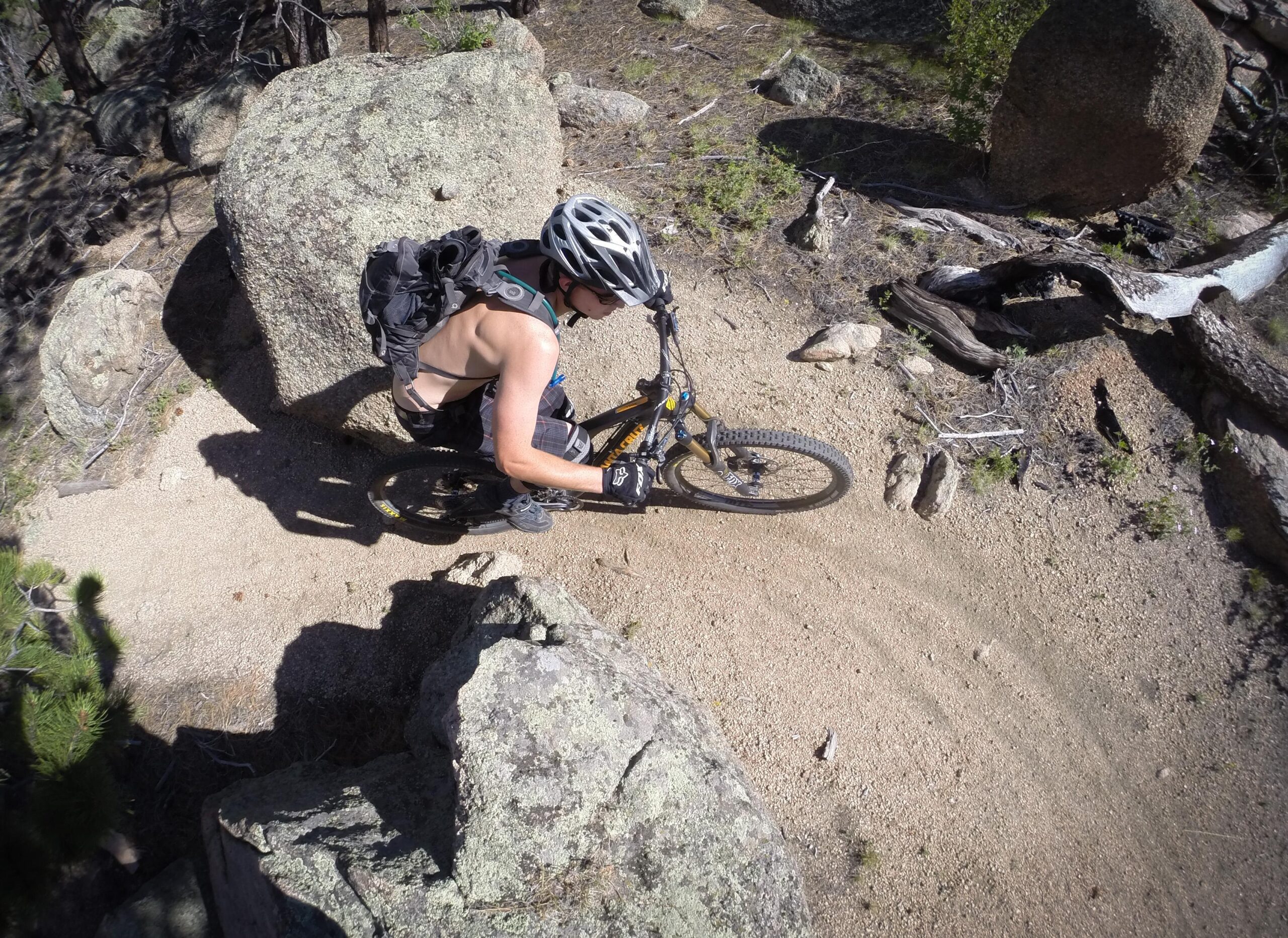 A mountain biker navigating a winding dirt trail surrounded by large rocks and trees. The rider is wearing a helmet and gloves, and has a backpack on their back. The image captures the action and excitement of mountain biking in a natural outdoor setting. Blackjack / Raspberry Ridge mountain bike trail.