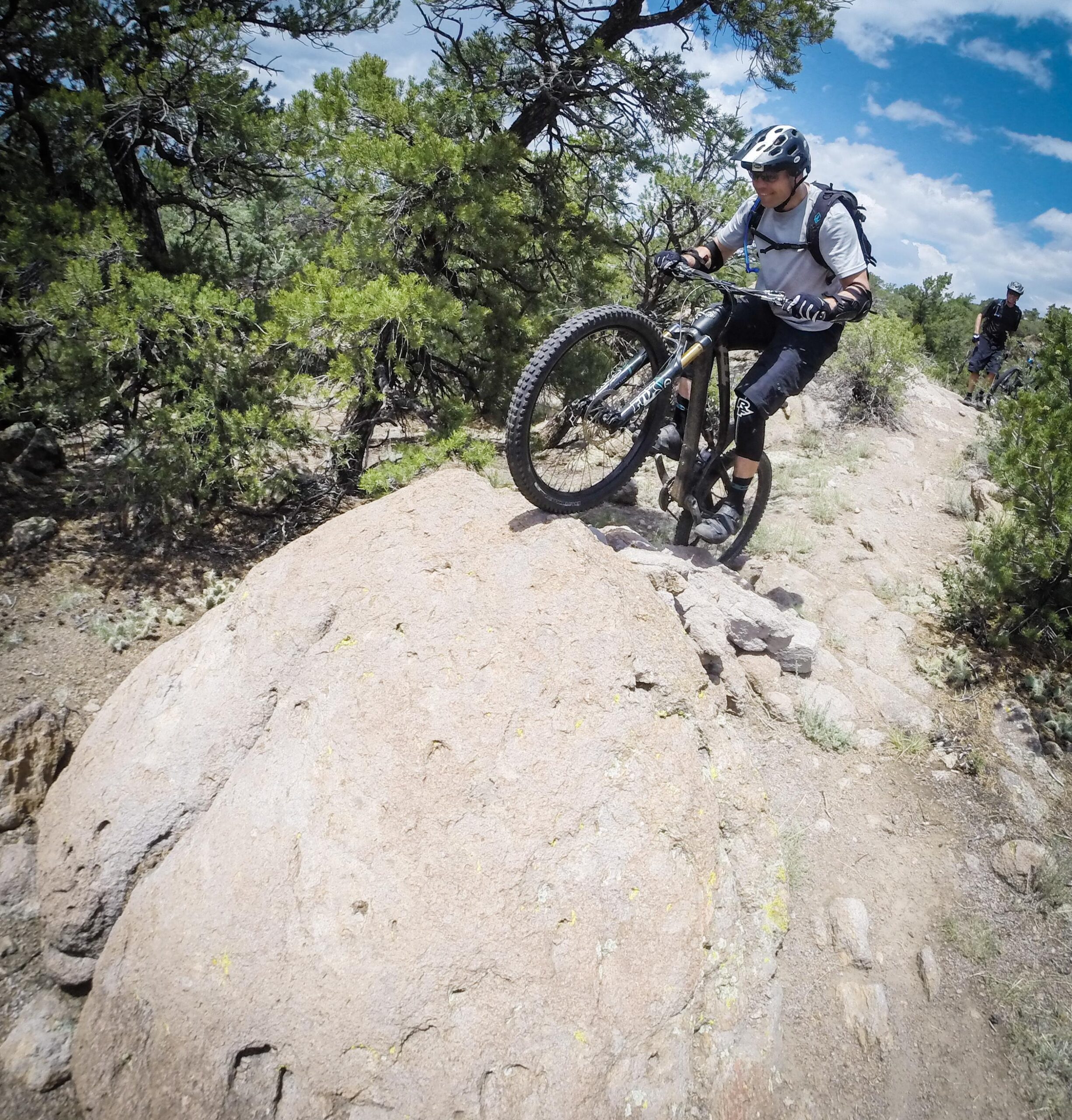 A mountain biker skillfully navigating a rocky terrain, lifting the front wheel while balancing on a large boulder, surrounded by trees and a clear blue sky. Arkansas Hills mountain bike trail.