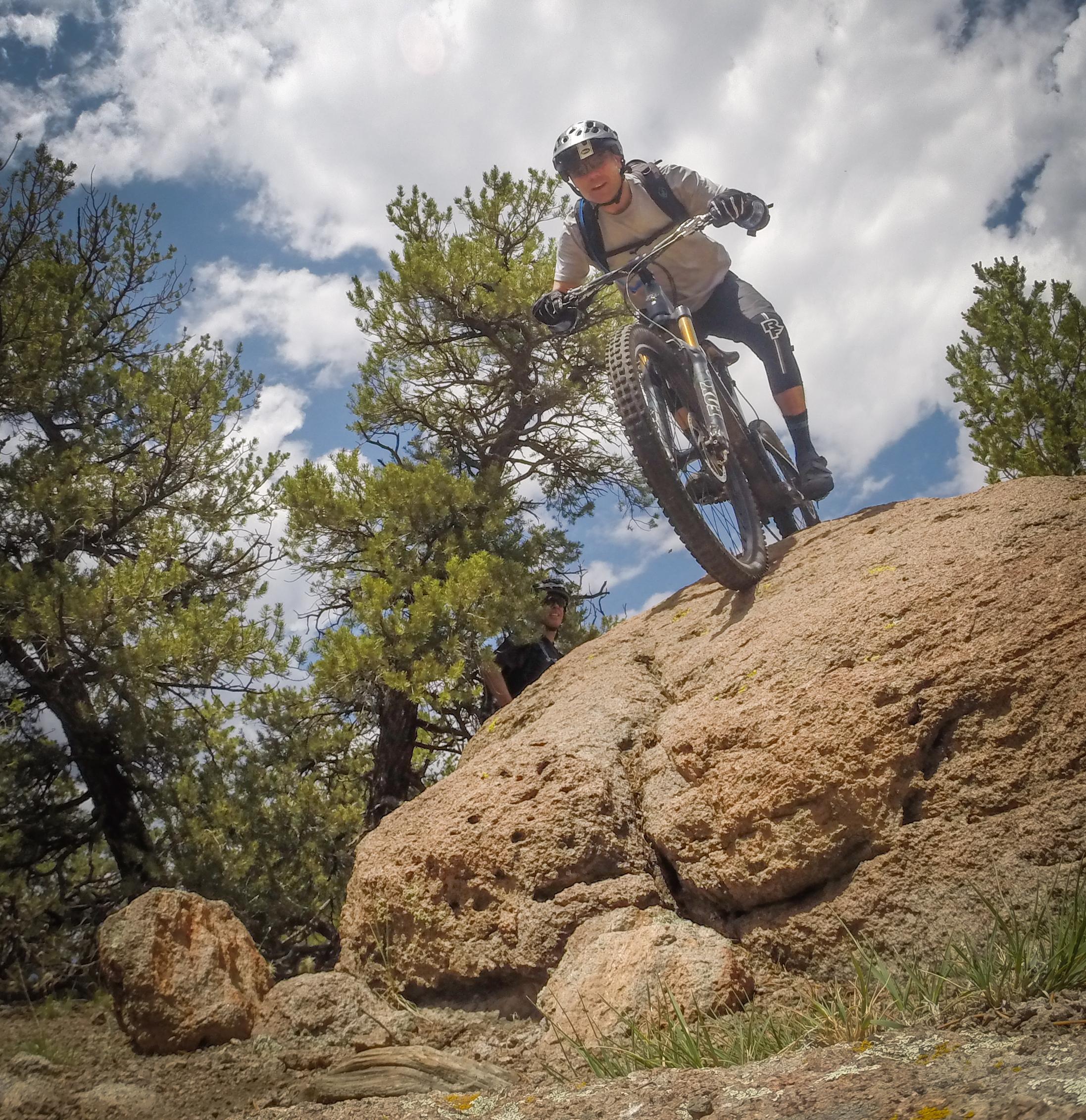 A mountain biker navigating a rocky trail, poised on a large boulder with trees and a blue sky in the background. Another cyclist can be seen in the distance, partially obscured behind the rock. Arkansas Hills mountain bike trail.