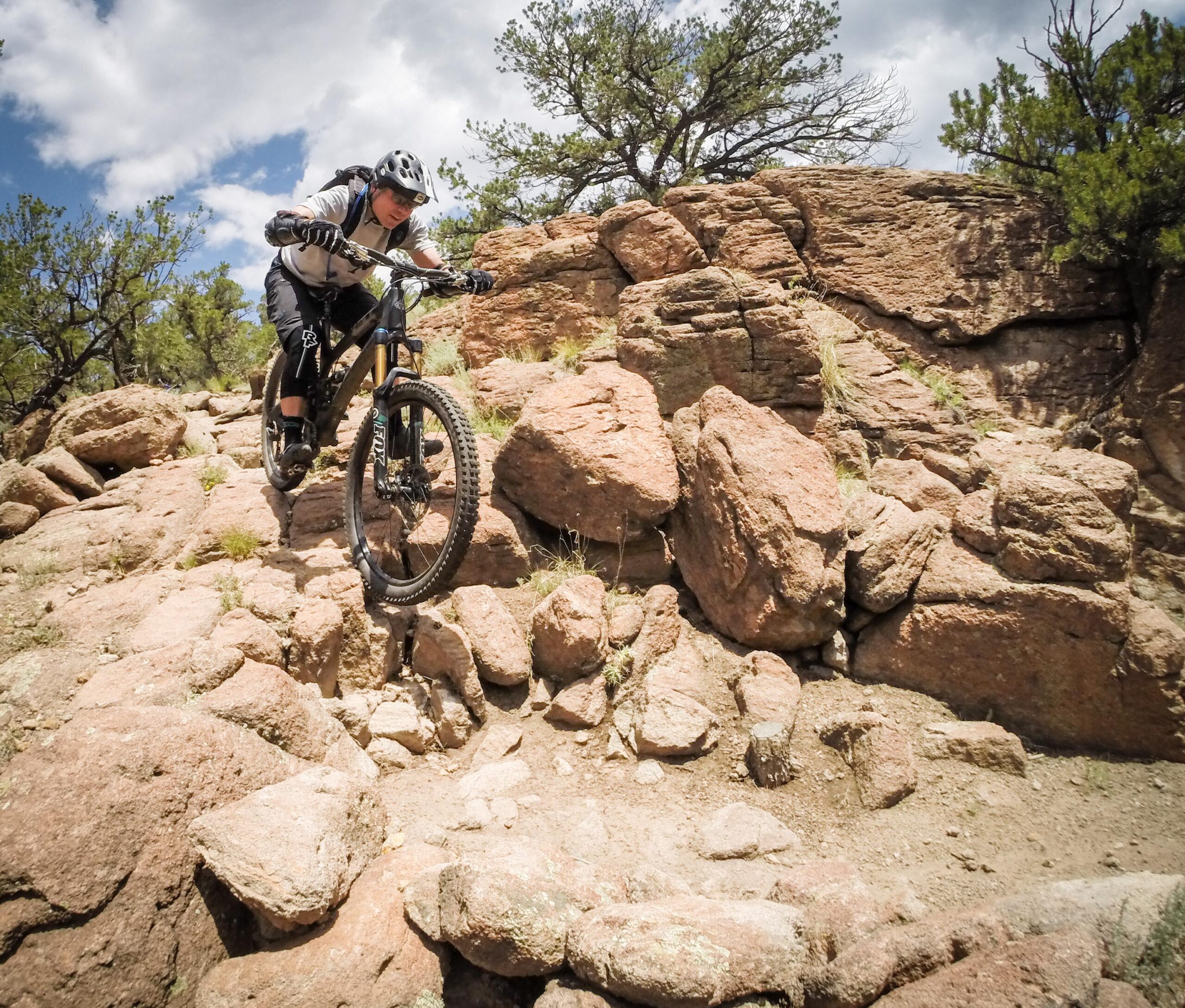 A mountain biker in protective gear navigates rocky terrain, poised to jump over a steep section of rocks, with trees and partly cloudy skies in the background. Arkansas Hills mountain bike trail.