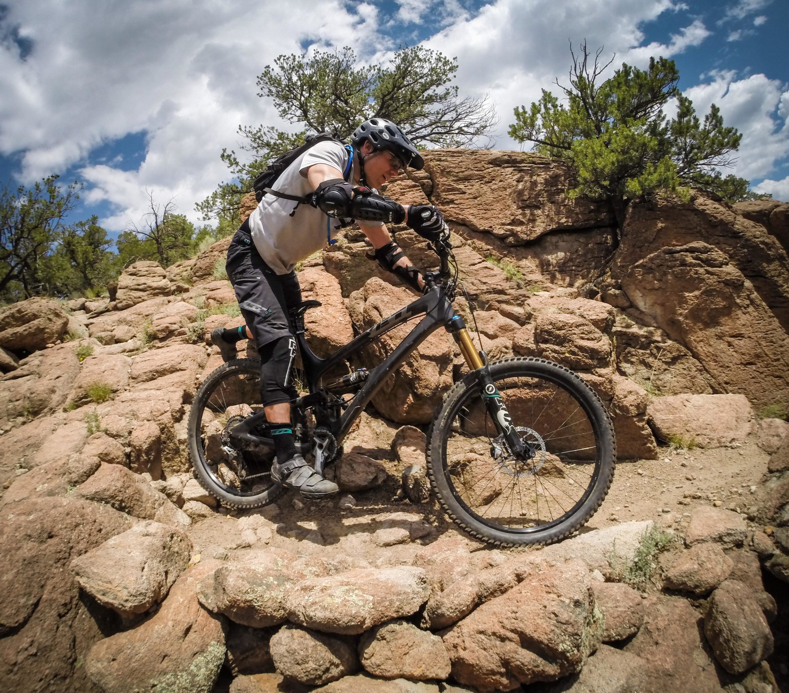 A mountain biker skillfully navigates a rocky trail, leaning forward on a black mountain bike as he climbs over large stones, under a partly cloudy sky filled with blue and white clouds. Surrounding him are sparse trees and rugged terrain, highlighting the challenging nature of the trail. Arkansas Hills mountain bike trail.