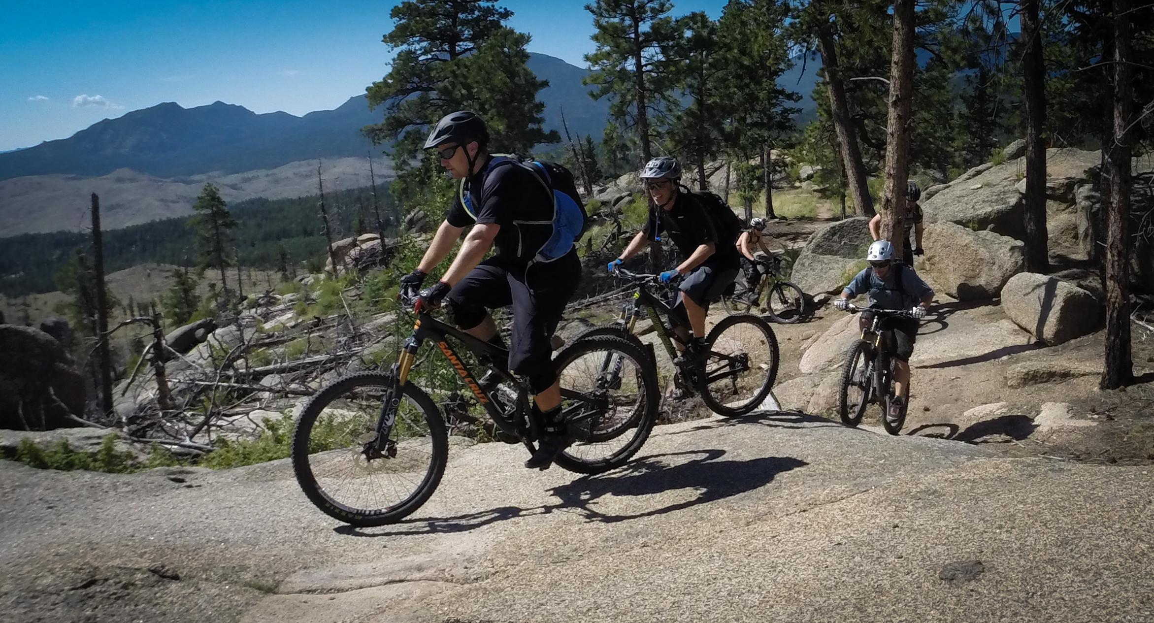 A group of mountain bikers navigating a rocky trail in a forested area, with mountains visible in the background. The sun is shining, and the terrain features boulders and sparse vegetation. Buffalo Creek mountain bike trail.