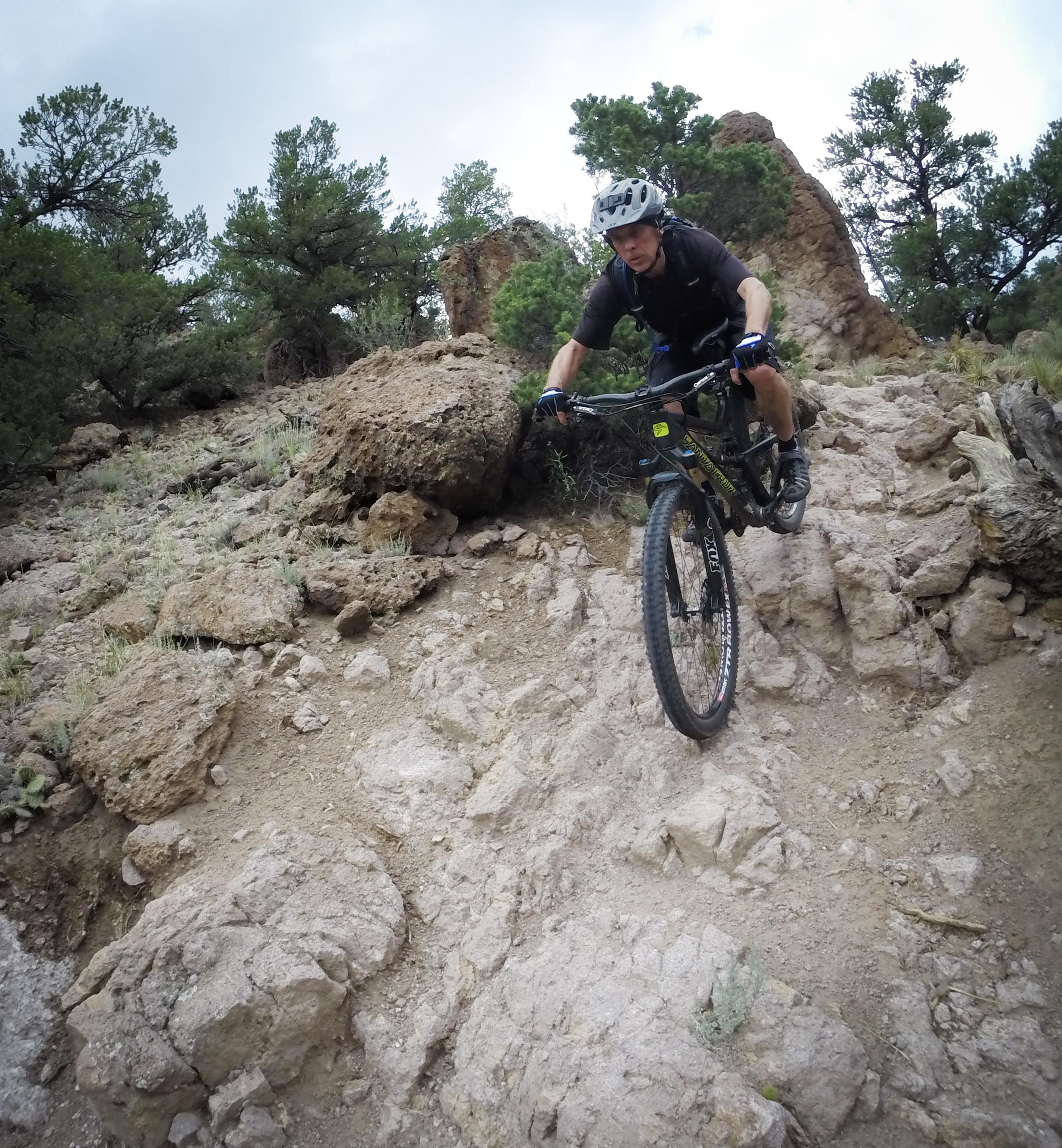 A person riding a mountain bike down a rocky trail surrounded by sparse vegetation and trees. The rider is wearing a helmet and biking gear, demonstrating a dynamic pose as they navigate the challenging terrain. Arkansas Hills mountain bike trail.
