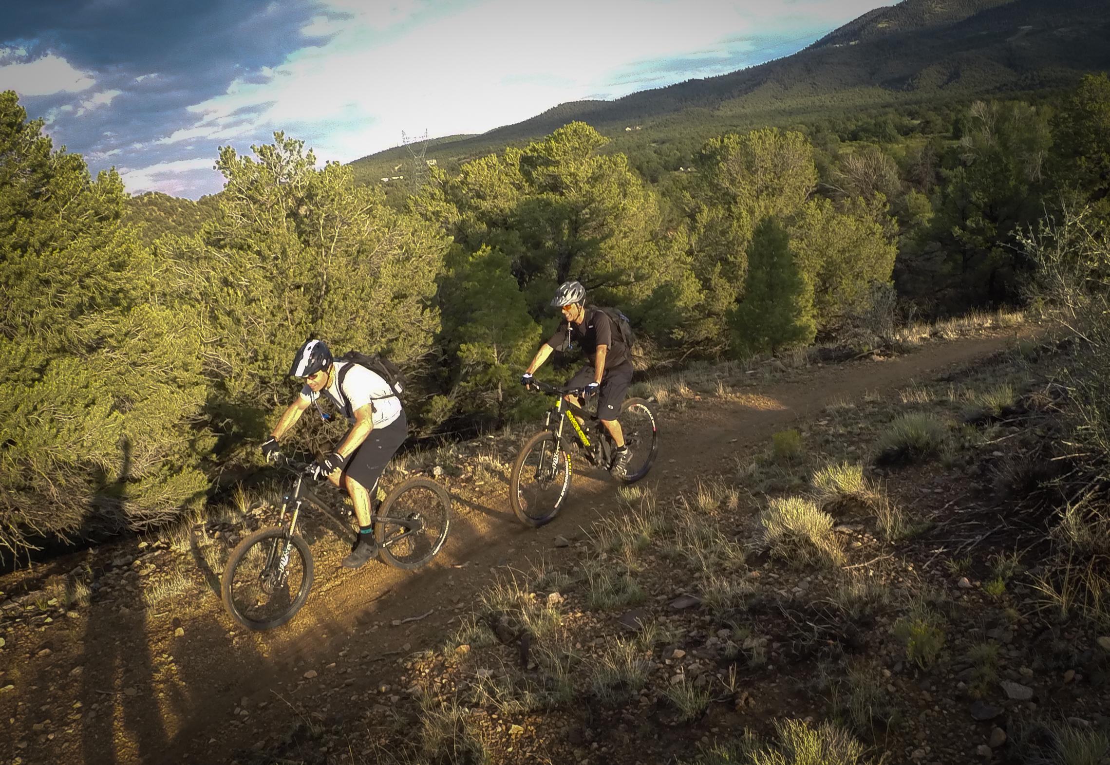 Two mountain bikers navigating a dirt trail surrounded by lush greenery and rolling hills under a partly cloudy sky. One rider is leaning forward on a black bike, while the other follows closely behind on a yellow bike, showcasing a scenic outdoor adventure. Methodist Mountain mountain bike trail.