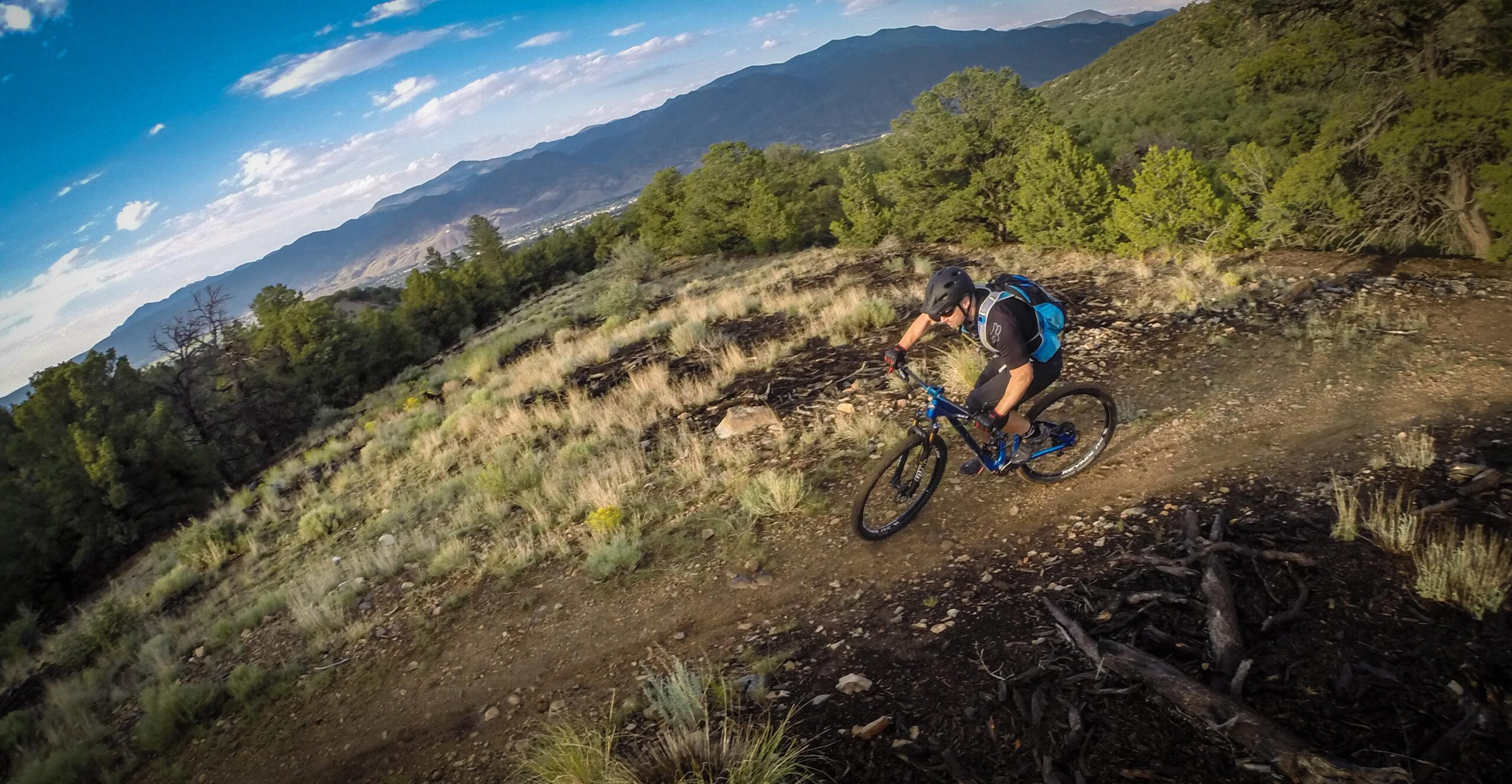 A mountain biker navigates a curvy dirt trail surrounded by greenery and rocky terrain, with mountains in the background under a blue sky with scattered clouds. Methodist Mountain mountain bike trail.
