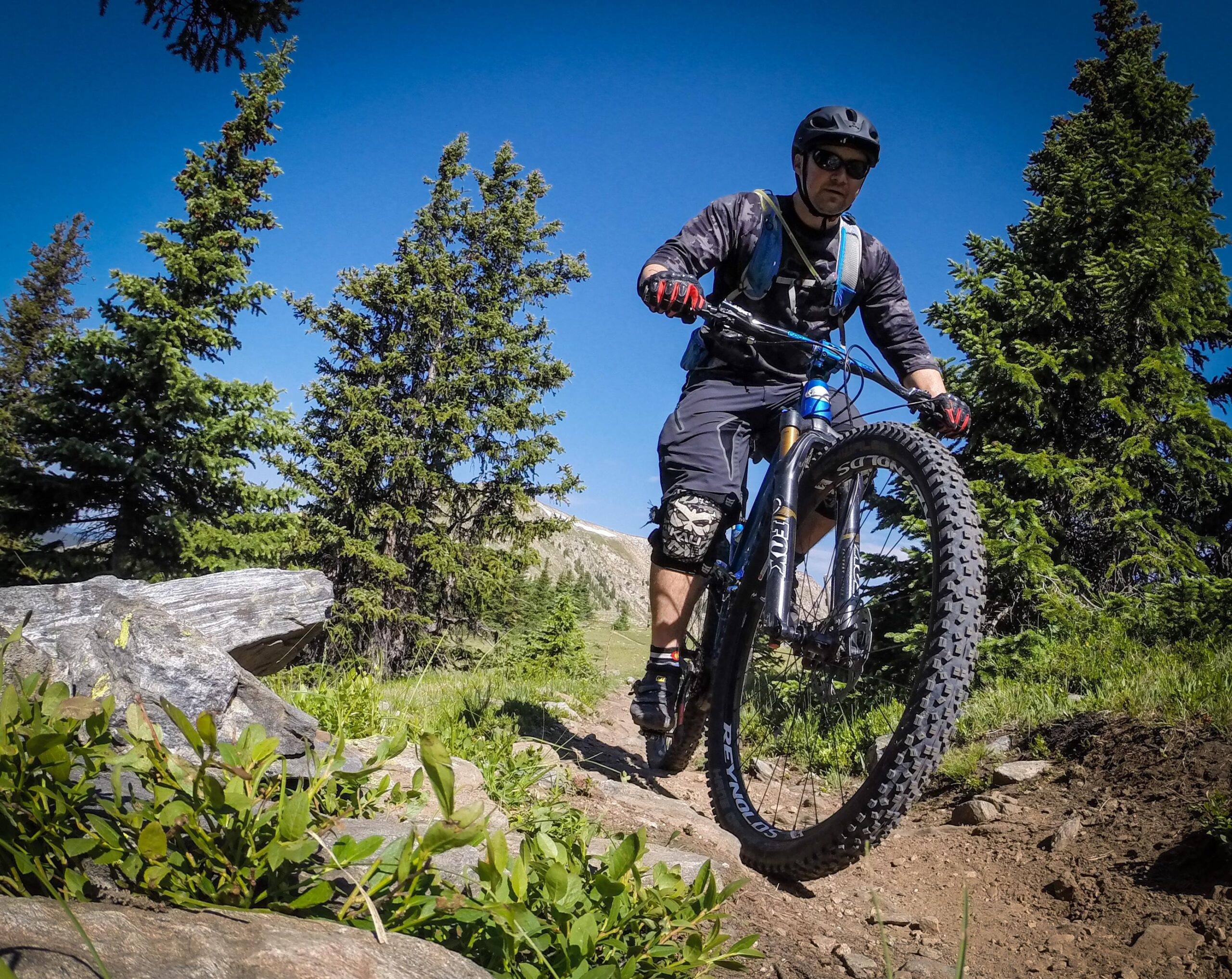 A mountain biker navigating a rocky trail surrounded by green pine trees under a clear blue sky. The cyclist is wearing a helmet, sunglasses, and protective gear, and is focused on maintaining balance as the bike's front wheel lifts slightly off the ground. Monarch Crest Trail mountain bike trail.