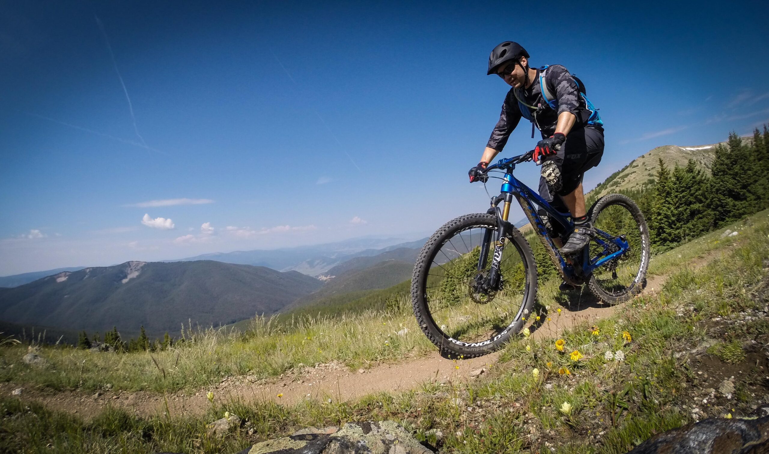 A mountain biker riding on a dirt trail through a grassy landscape, with rolling hills and mountains in the background. The sky is blue with a few clouds, and wildflowers can be seen along the trail. The cyclist is wearing a helmet and riding gear, focused on the path ahead. Monarch Crest Trail mountain bike trail.