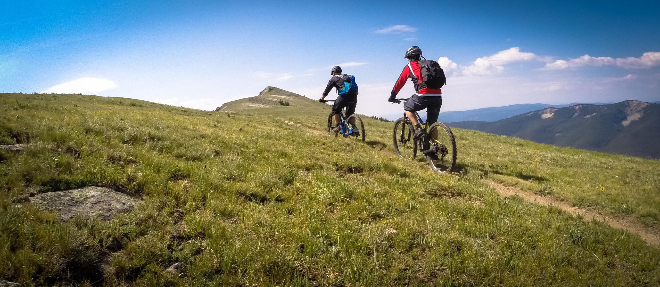 Two mountain bikers riding along a grassy trail on a hillside, with a blue sky and distant mountains in the background. The cyclists are wearing helmets and backpacks, and the landscape features lush green grasses and scattered rocks. Monarch Crest Trail mountain bike trail.