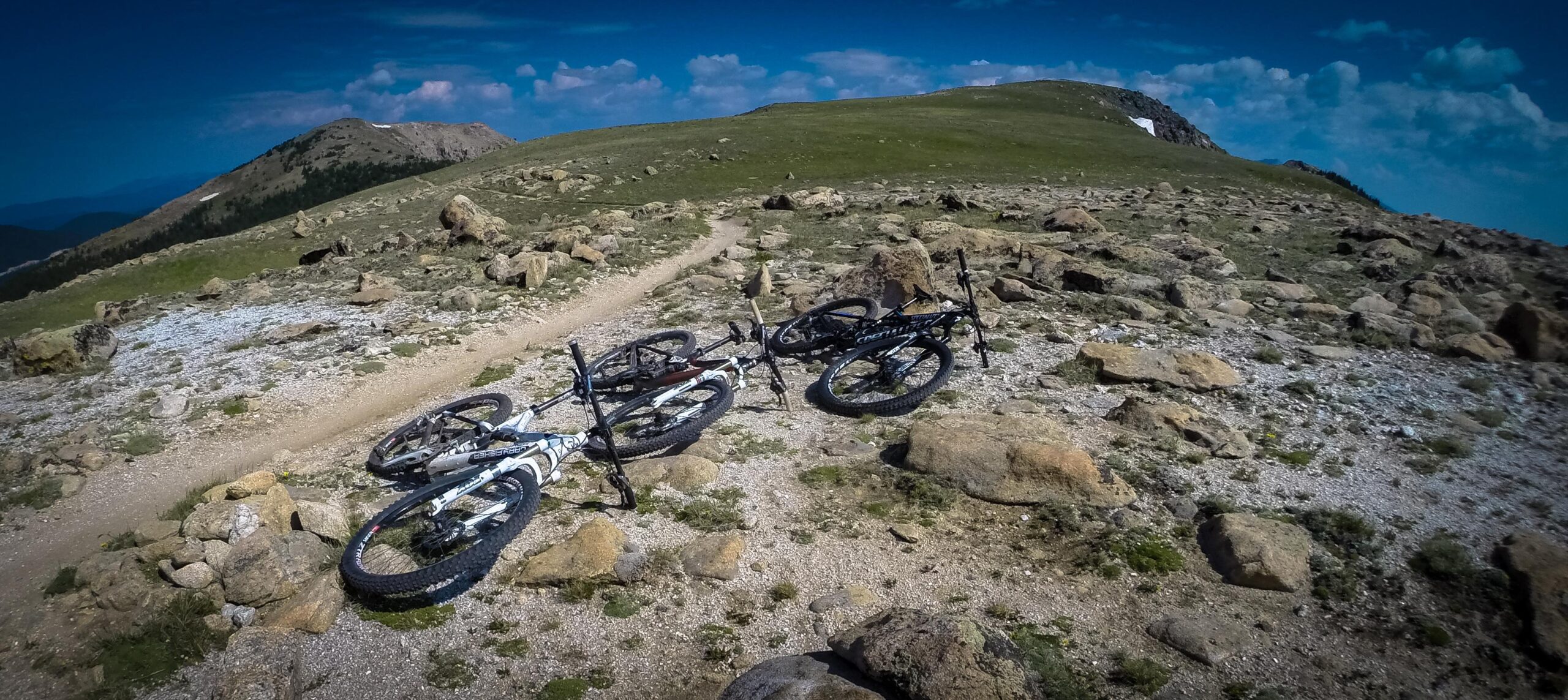 Alt tag: "Four mountain bikes lying on a rocky trail in a mountainous landscape with grassy areas and blue skies." Monarch Crest Trail mountain bike trail.