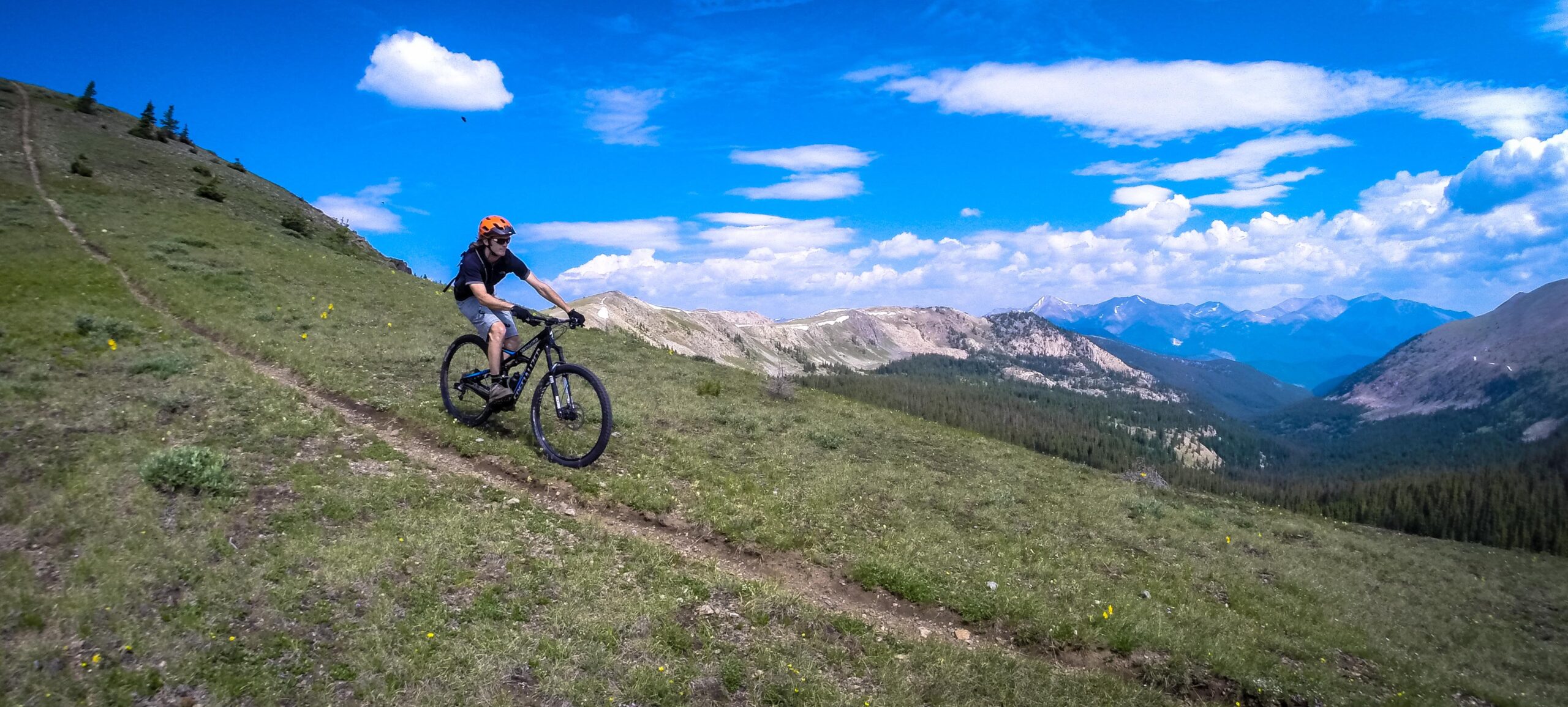 A mountain biker riding along a grassy terrain on a hillside, with distant mountains and a blue sky dotted with clouds in the background. Monarch Crest Trail mountain bike trail.